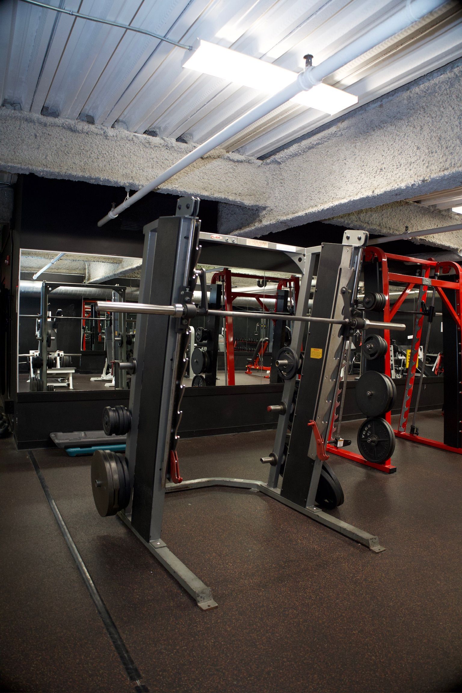 A Smith machine in a gym setting. The machine is silver with a barbell resting on it. The floor is black, and there are mirrors in the background.