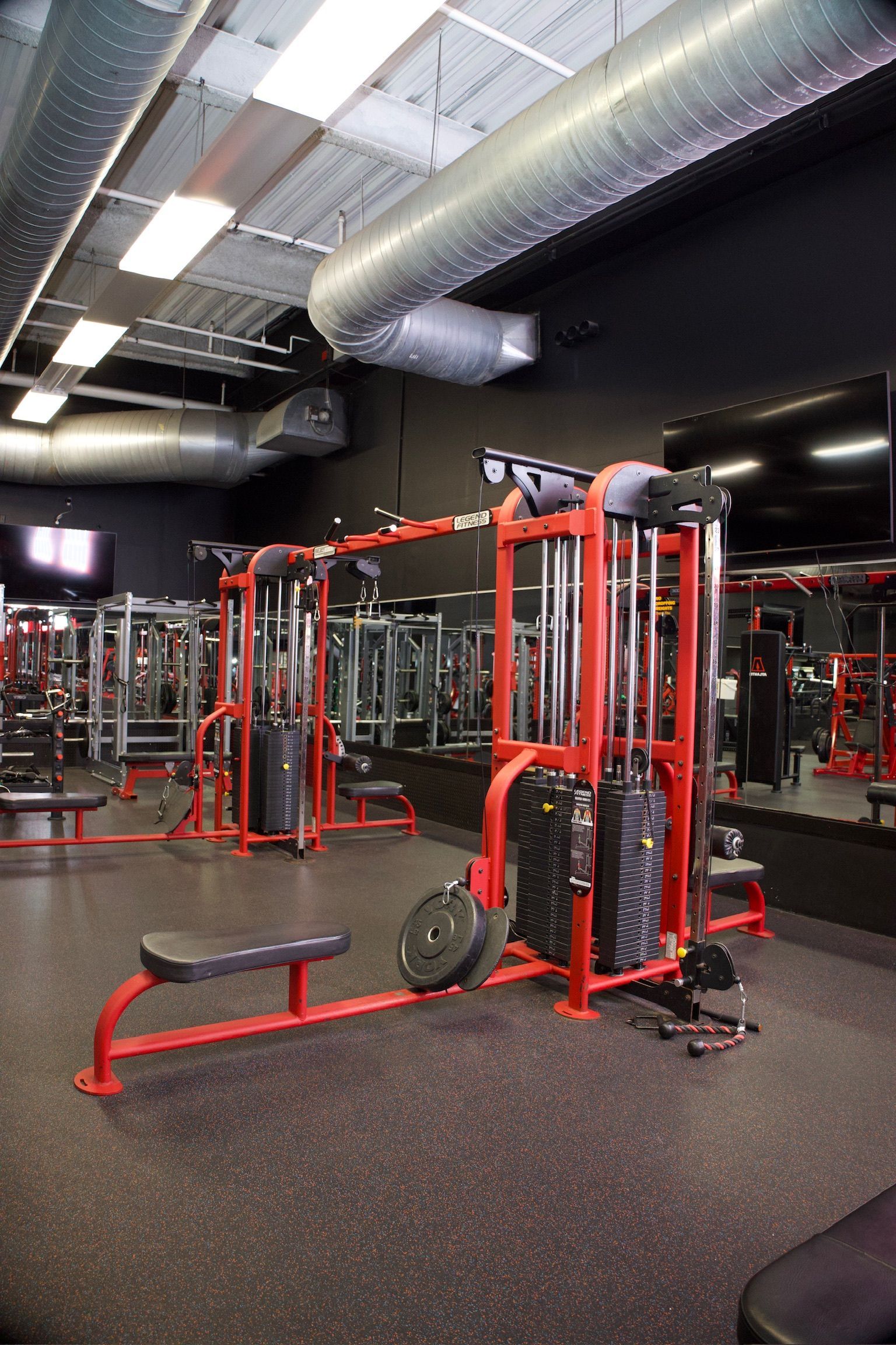 Interior of a gym with red and black equipment. A cable machine is in the foreground, with a mirrored wall behind.