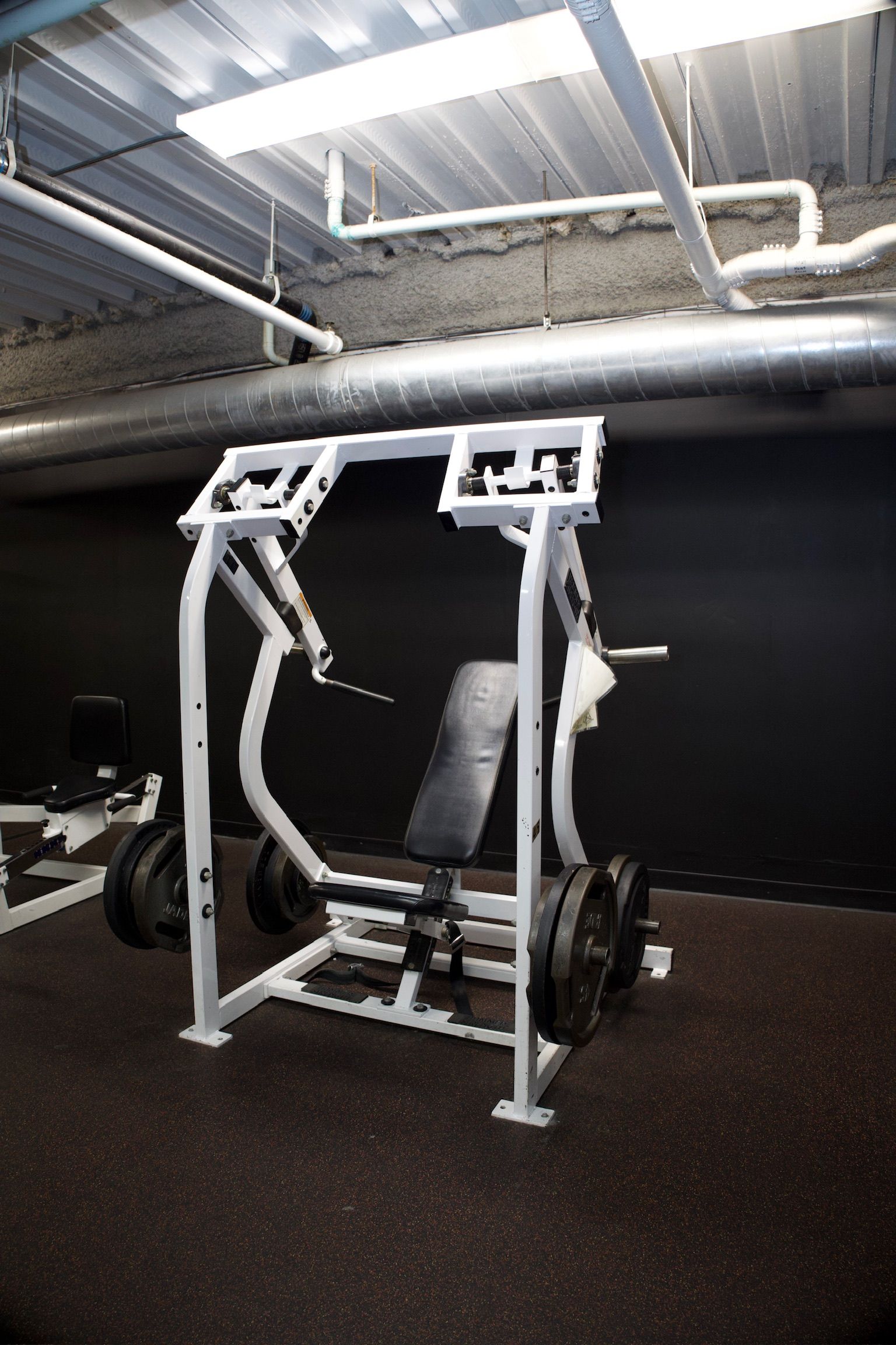 A weight machine in a gym. White frame, black weights, and seat. Dark flooring and walls.
