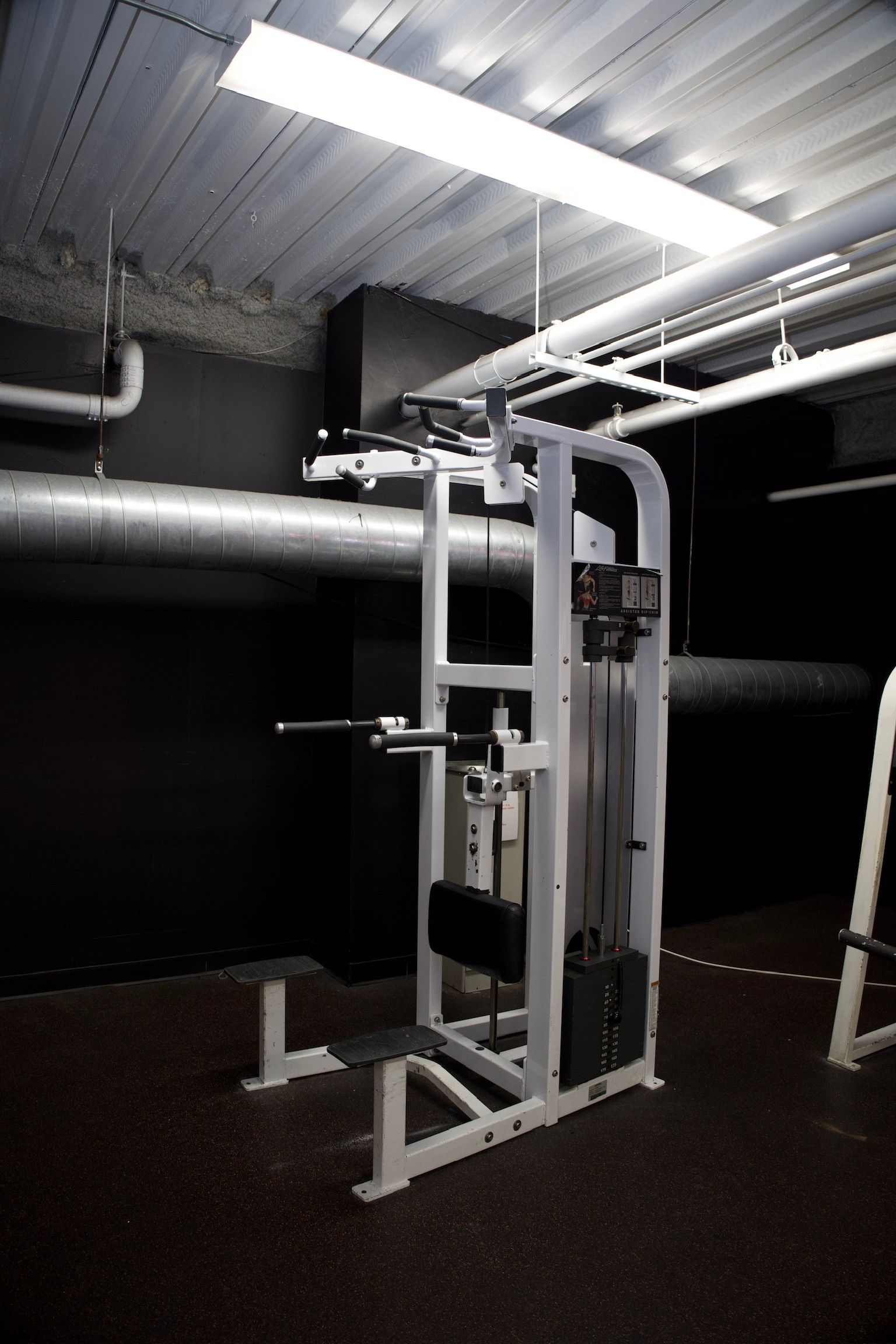 Weight machine in a dark gym setting. Features a pull-up bar, weights, and a seat.