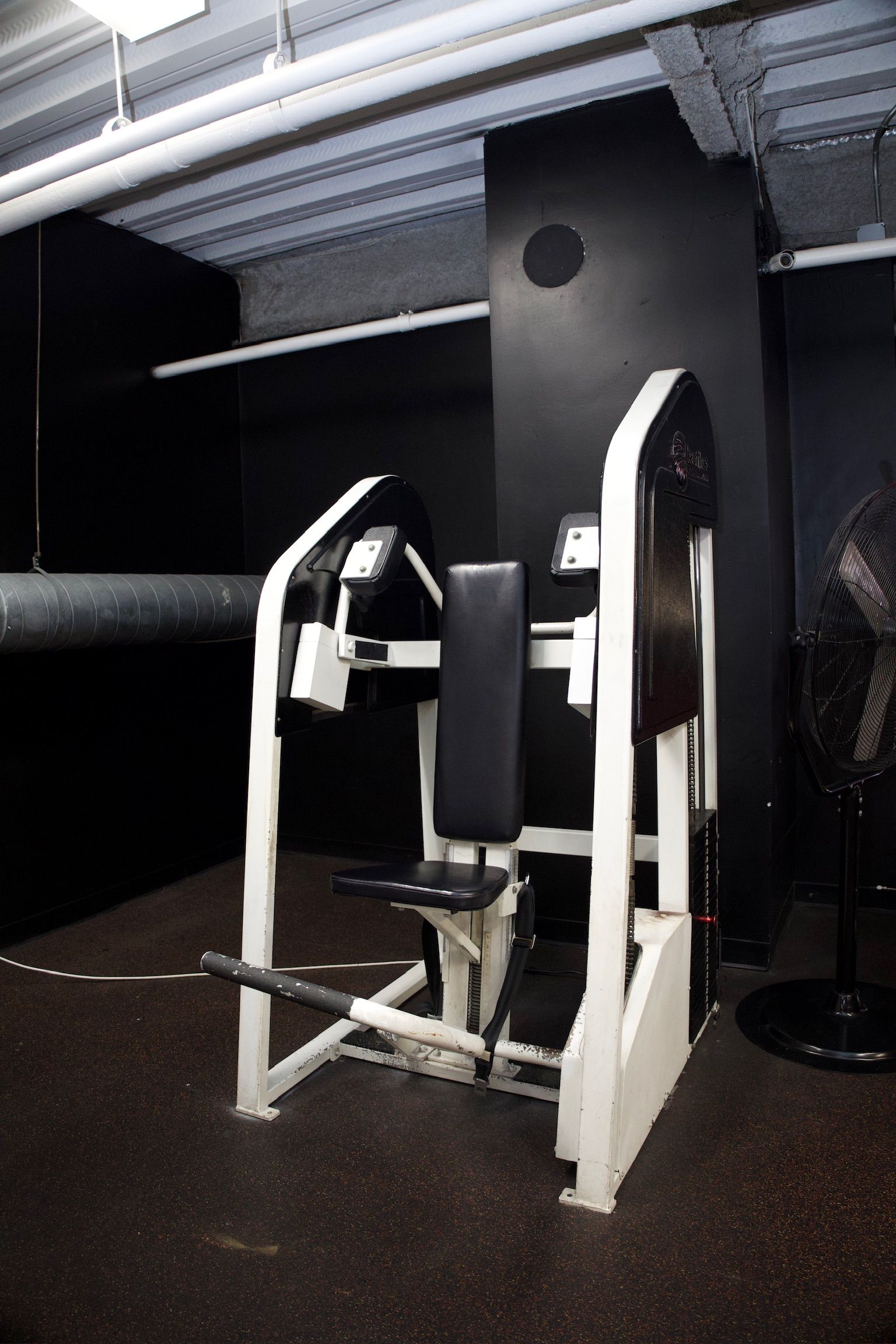A weight machine in a black-walled gym with a black seat and white frame.