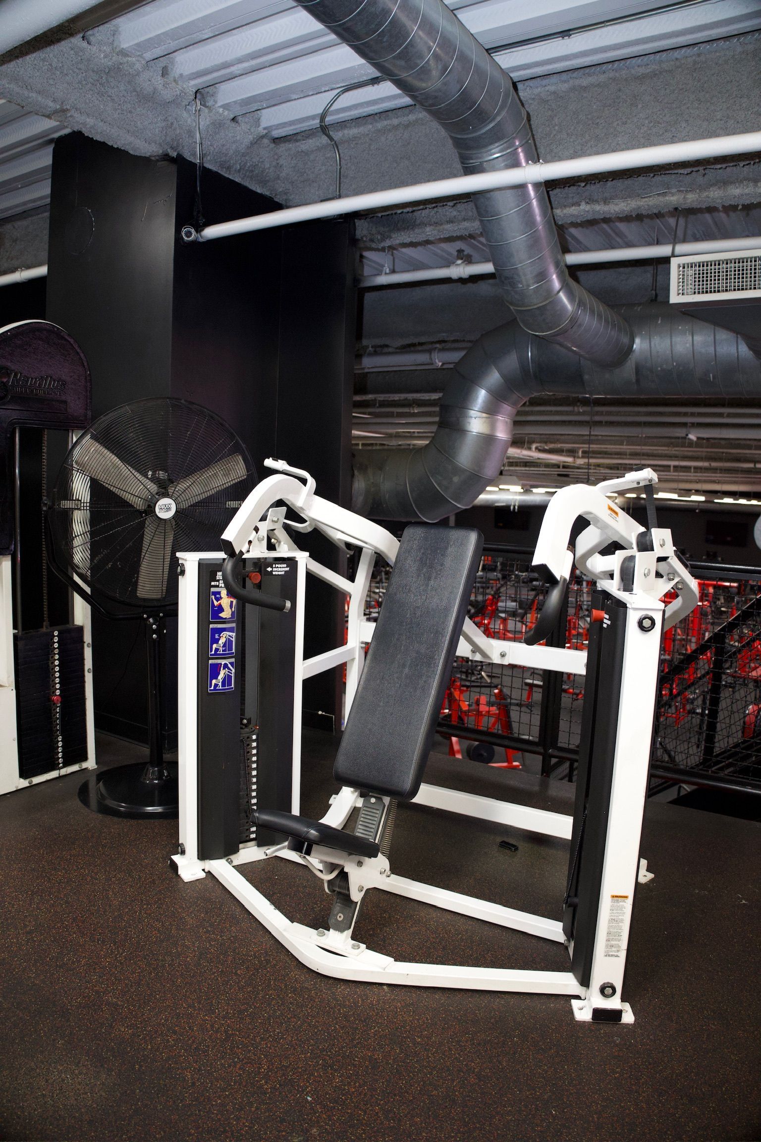Weight machine in a gym, angled view. White frame, black pad and accents, ventilation duct above.