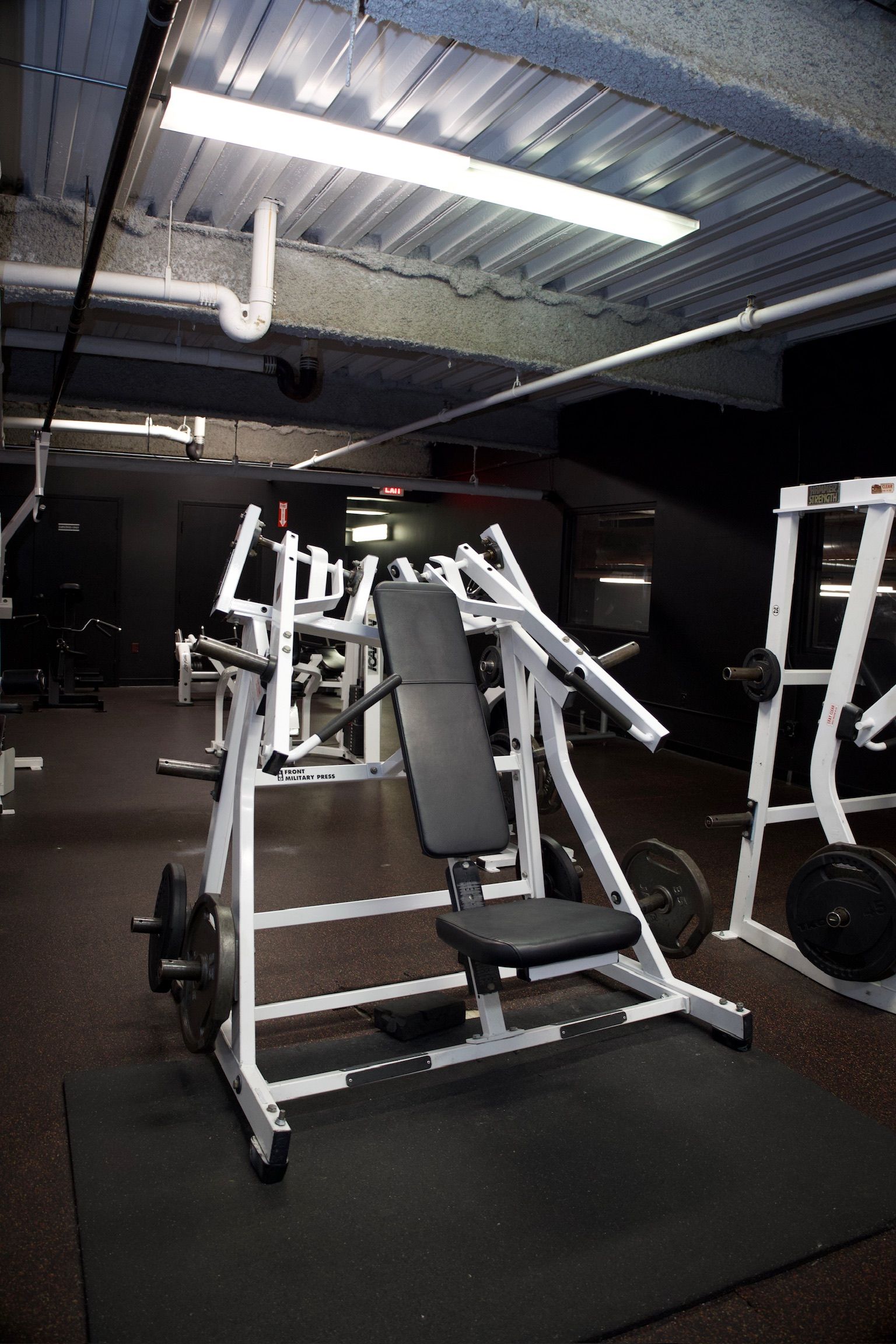 Weight machines in a dimly lit gym. A white chest press machine is in the foreground, with black weight plates.
