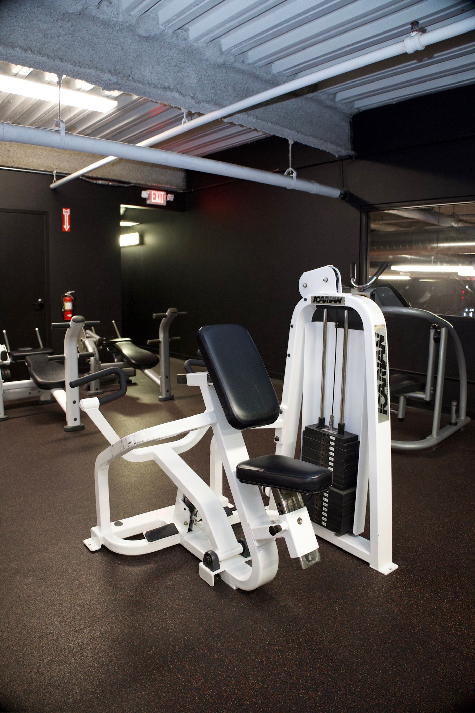 Gym equipment in a fitness center. White weight machine with black seat, black floor, and dark gray walls.