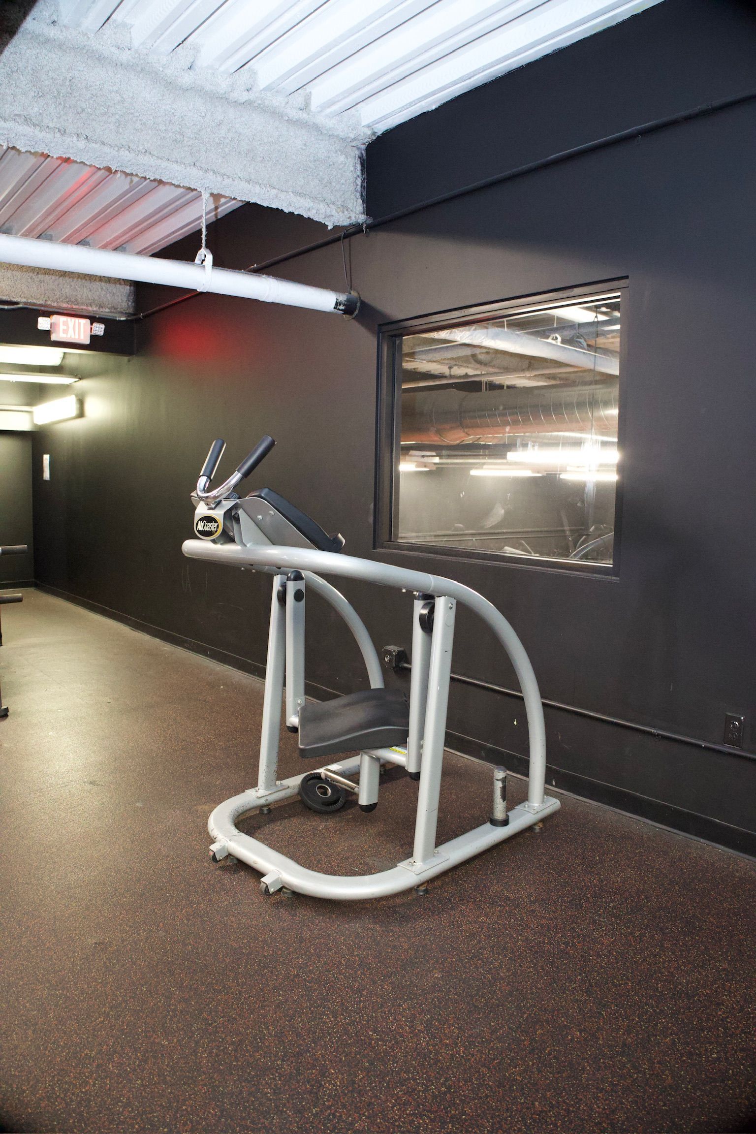 Gym interior: Exercise machine against a dark wall. The window reflects a bright area. Red rubber flooring.