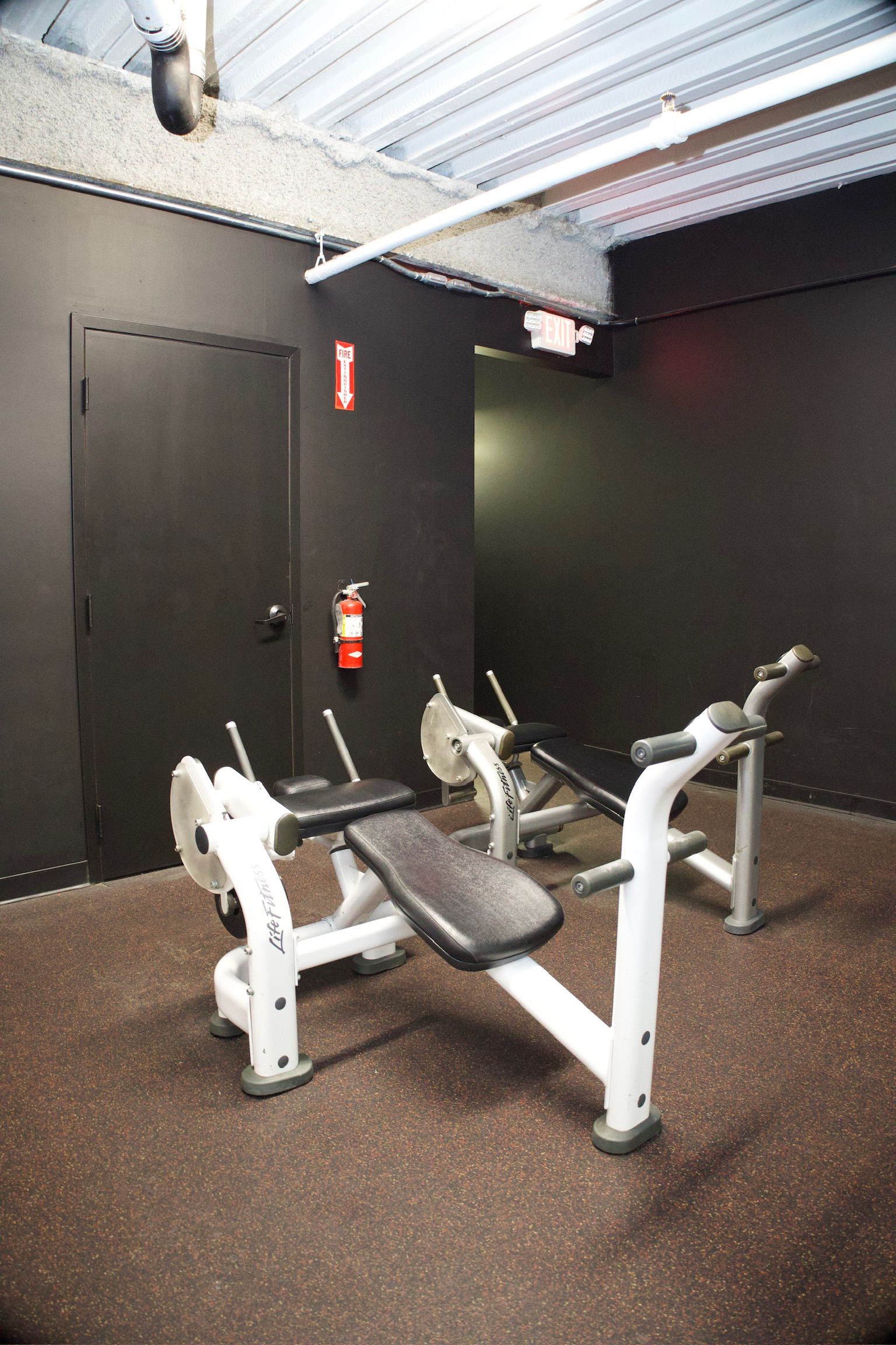 Gym interior with white weight machines against dark walls, a fire extinguisher, and a door.