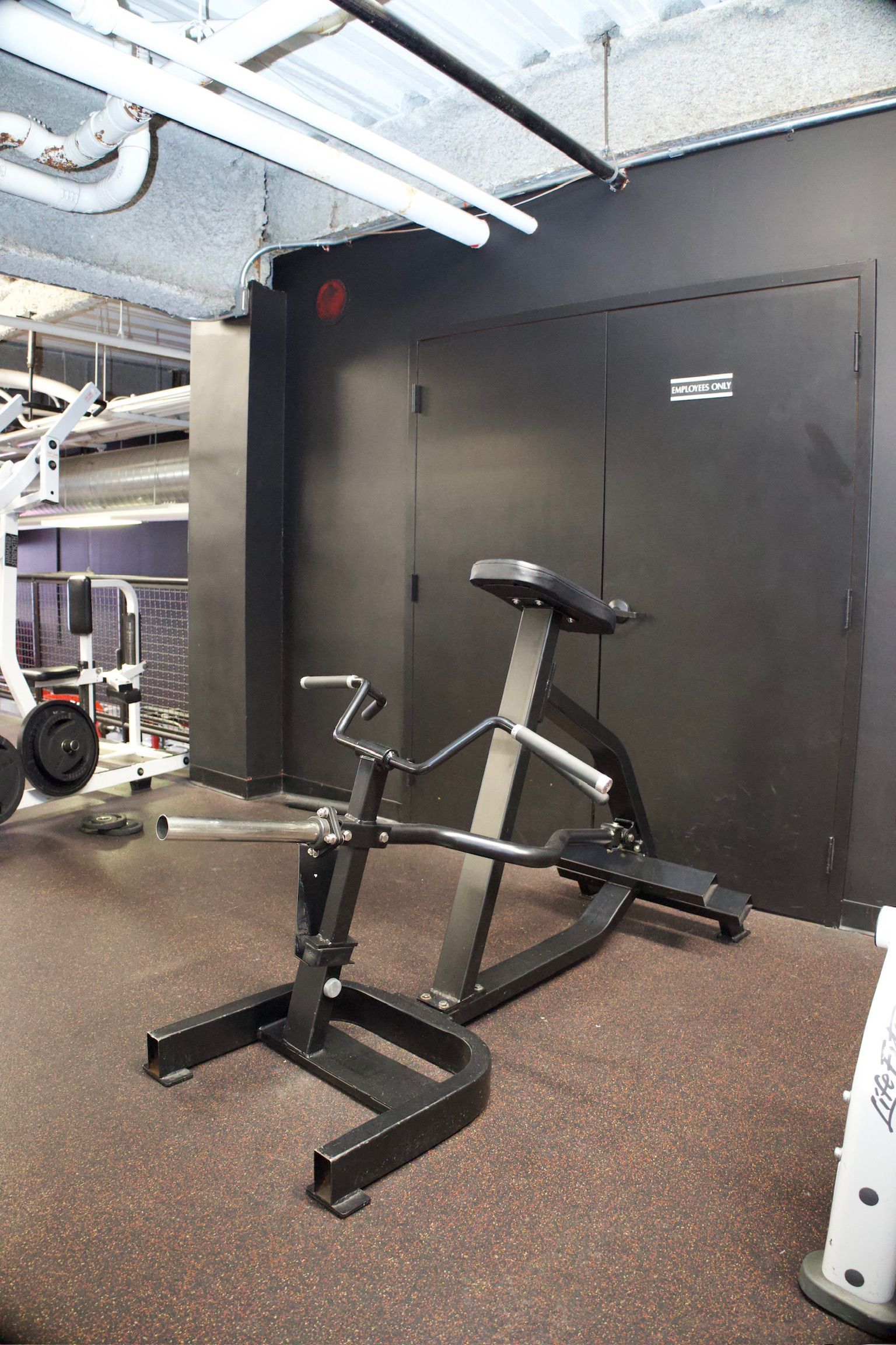 Weightlifting machine in a gym setting. Black frame, angled back support, and barbell holder on a brown floor.