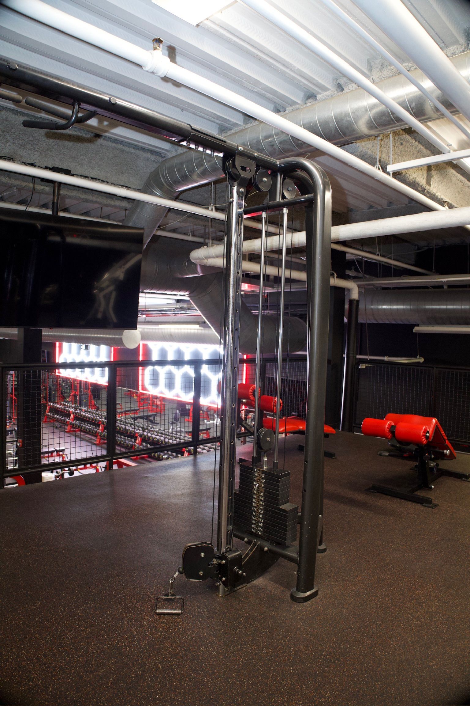 A cable machine in a gym setting. Black metal frame with weights, red accents, and black rubber flooring.