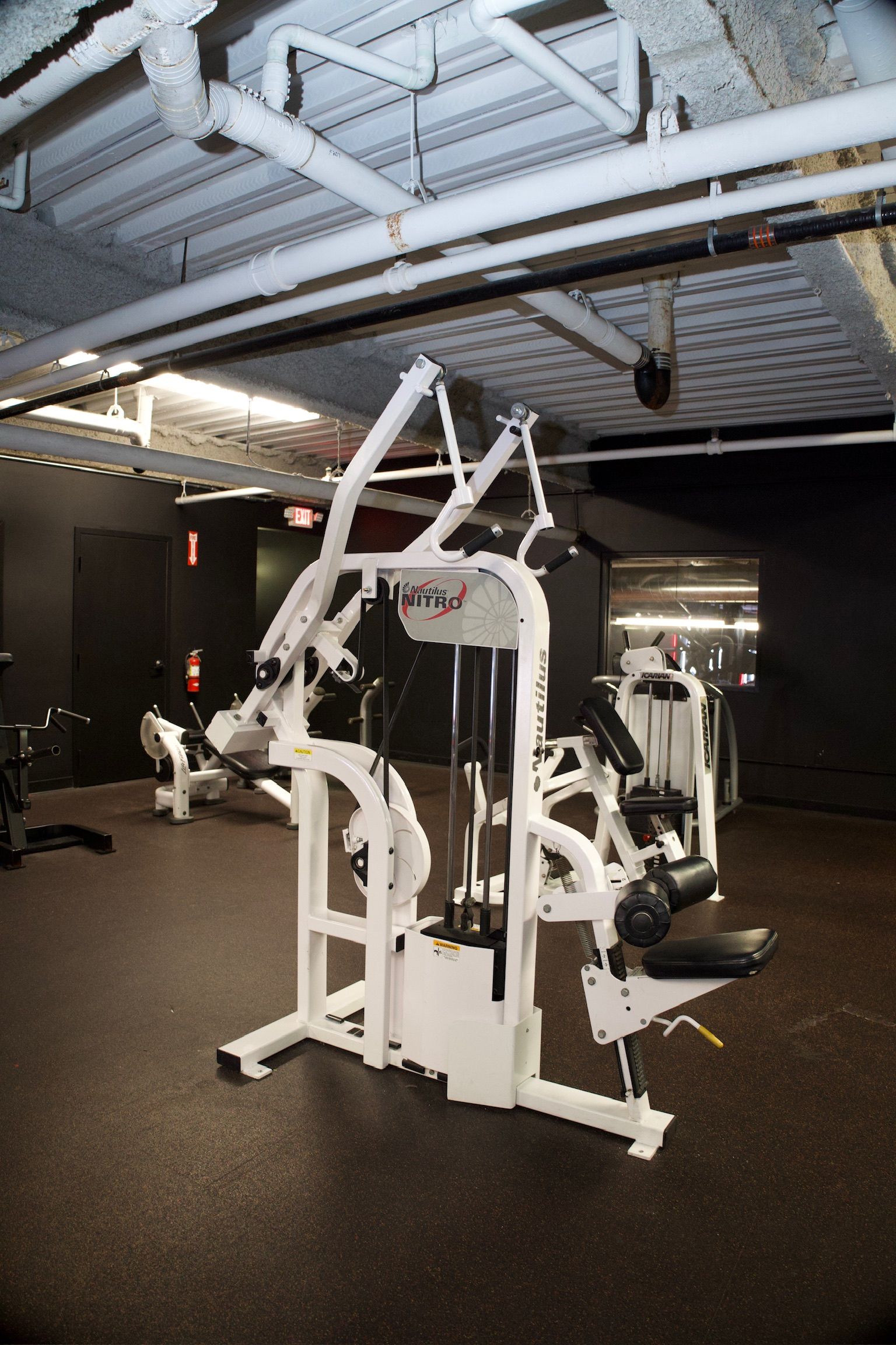 A white weight machine in a gym. Black walls and ceiling with exposed pipes.