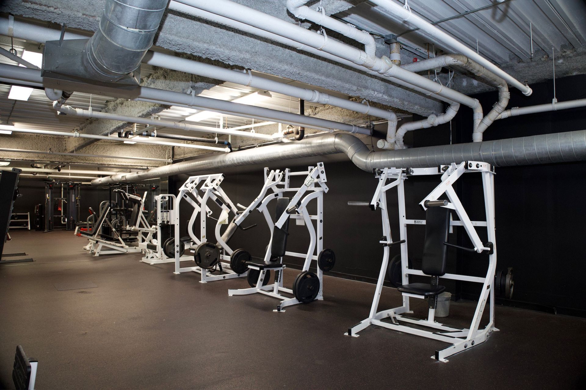Gym with weight machines and ventilation ducts. Black walls, dark floor, and white exercise equipment.