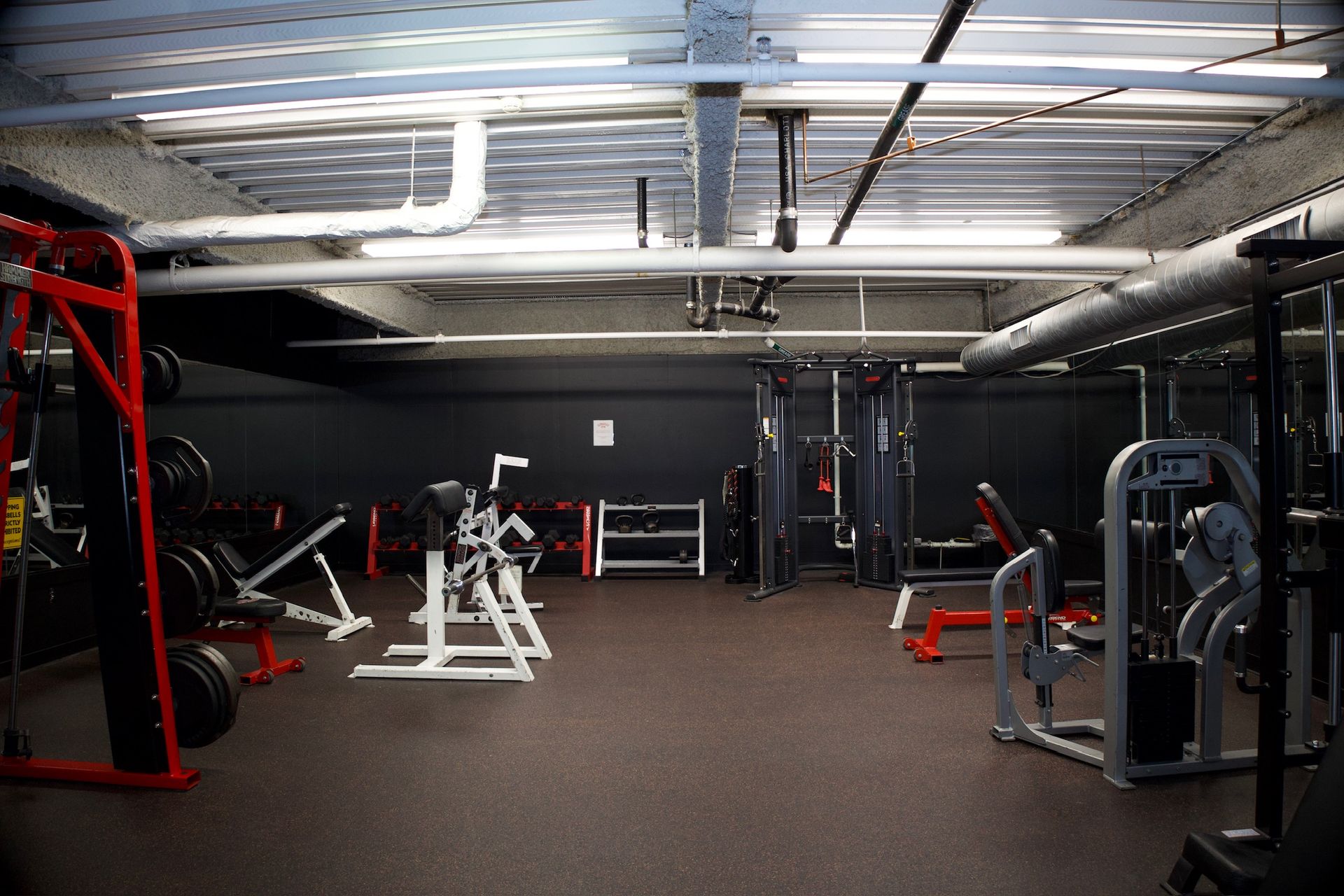 Weight room with weight machines and free weights. Dark walls, metal beams, and a rubber floor.