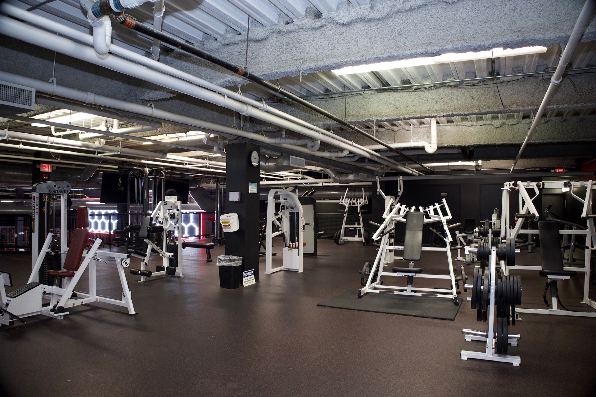 Weight machines in a dimly lit gym with black flooring and white support beams on the ceiling.