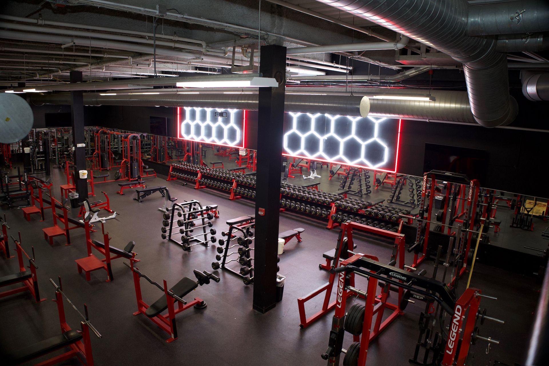 Interior view of a modern gym with red and black weightlifting equipment. The space has a high ceiling and neon lighting.