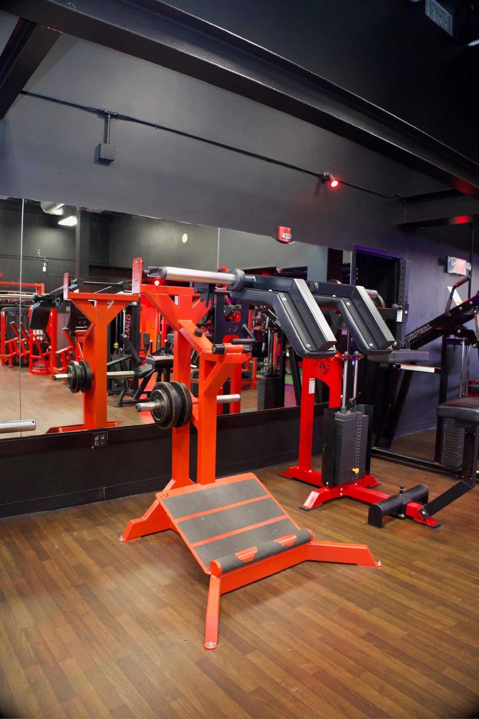 Red weight machines in a gym with wooden floors and a large mirror.