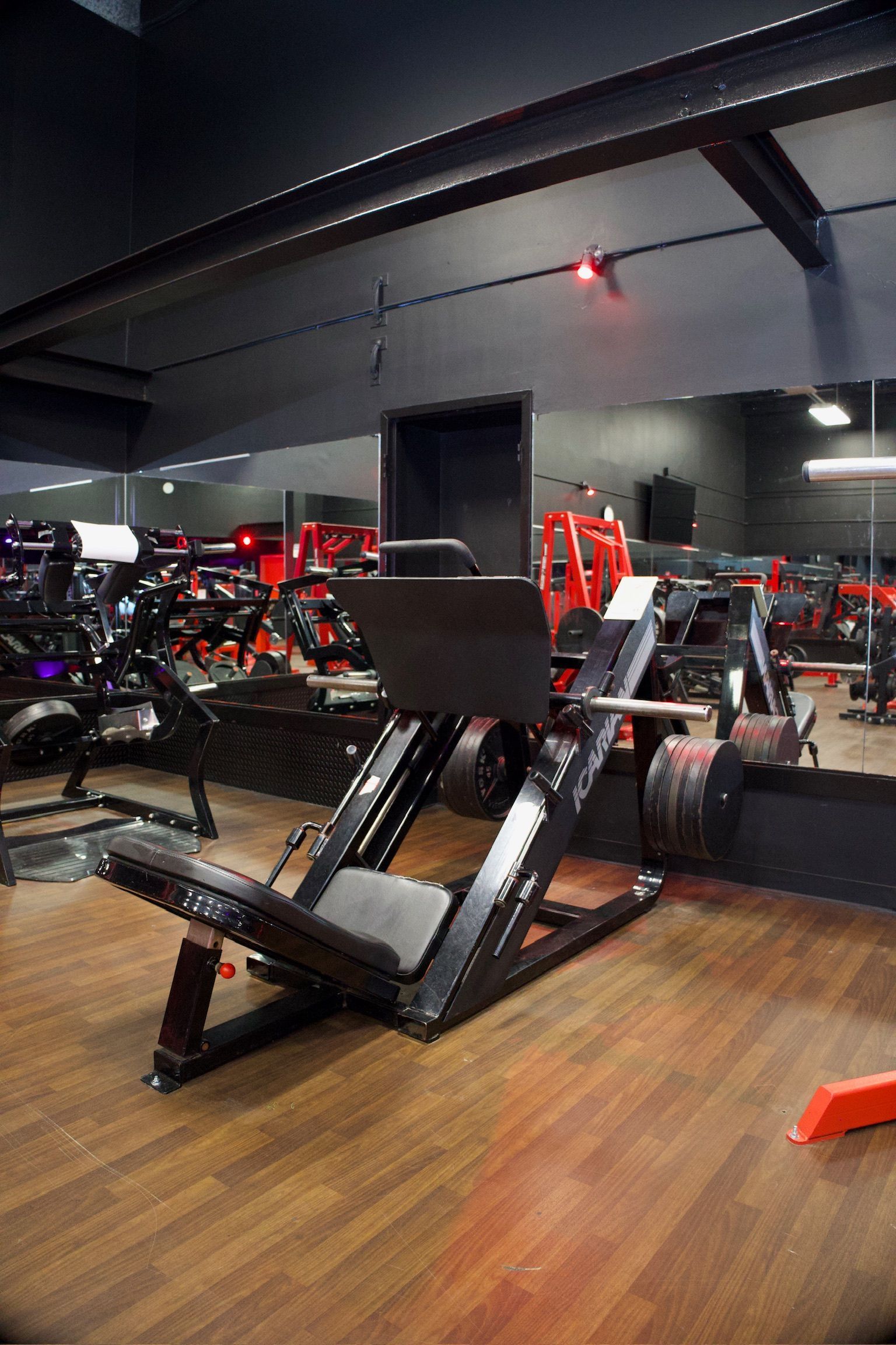 Leg press machine in a modern gym. Black and red interior, wooden floor, and weights.