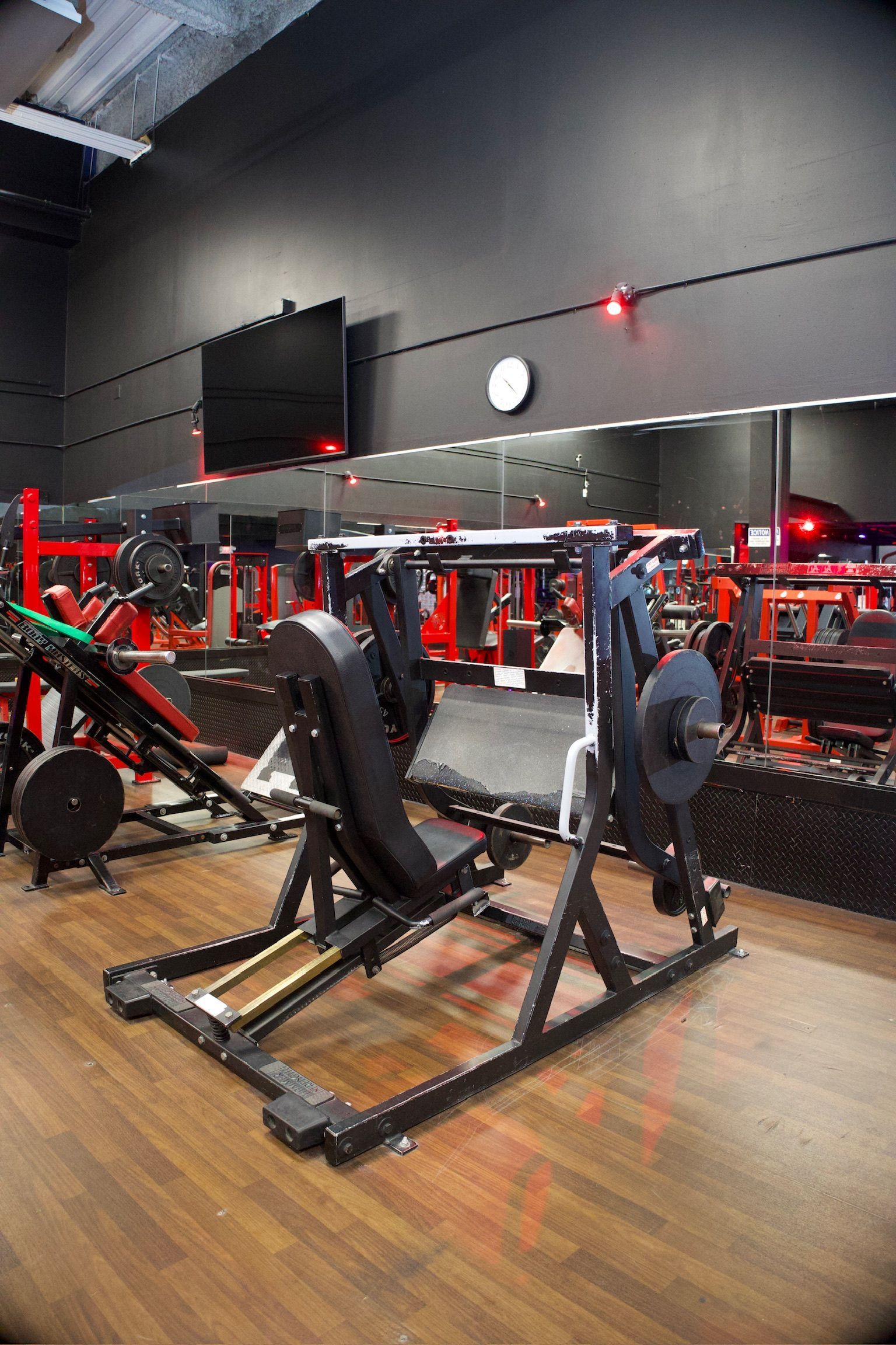 A weight machine in a gym. Black and red equipment on a wooden floor, black wall, and a mirror.
