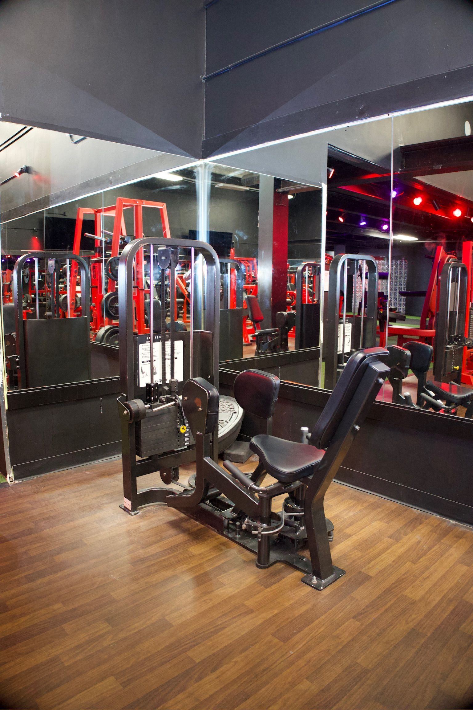 A weight machine in a gym with red accents and a wooden floor, reflected in a large mirror.