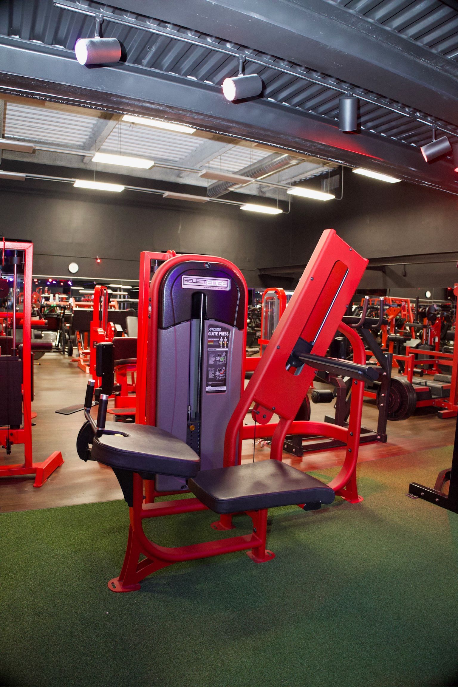 Red and black weight machine in a gym setting with green turf flooring.