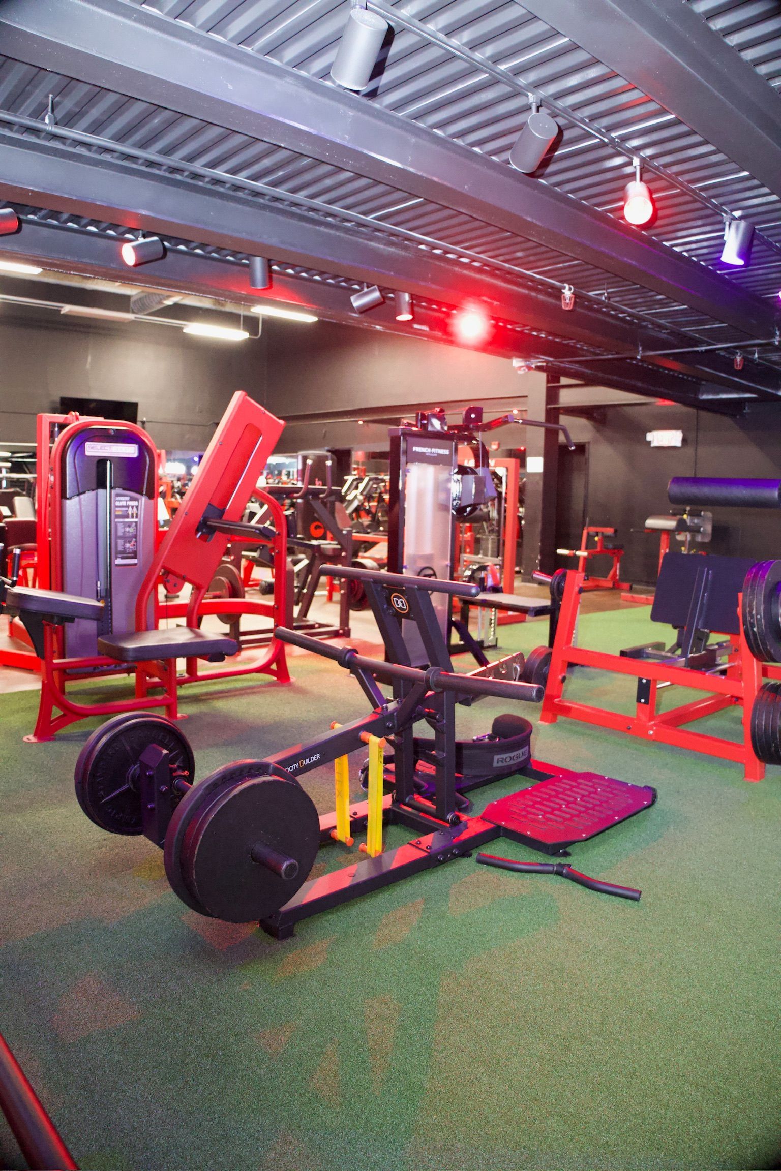 Weight machines in a gym with red and black equipment, green flooring, and red and purple lighting.