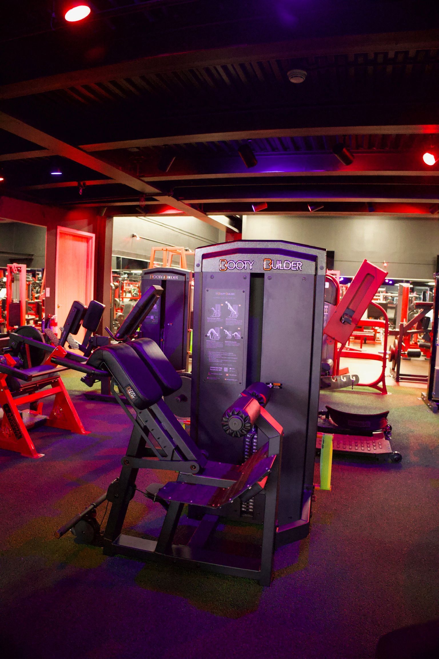 Gym interior with fitness machines and equipment, lit by red and purple lights.