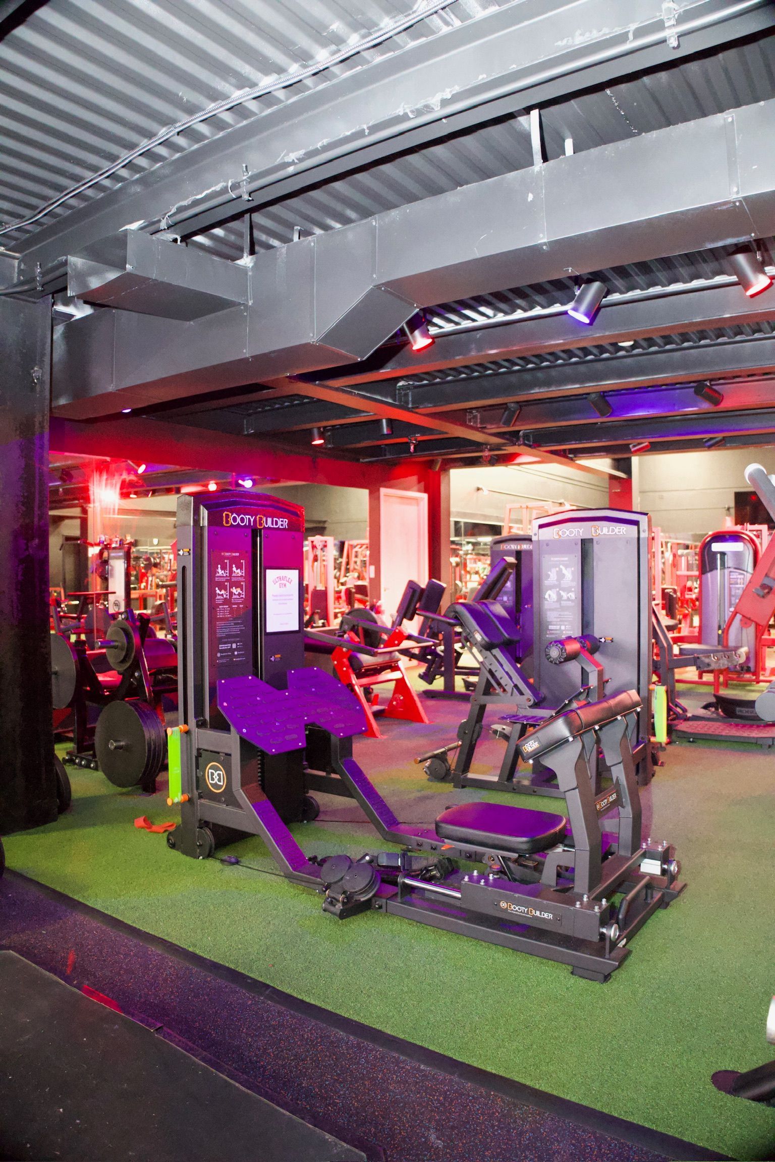 Gym interior with exercise machines on artificial turf under red and purple lighting.