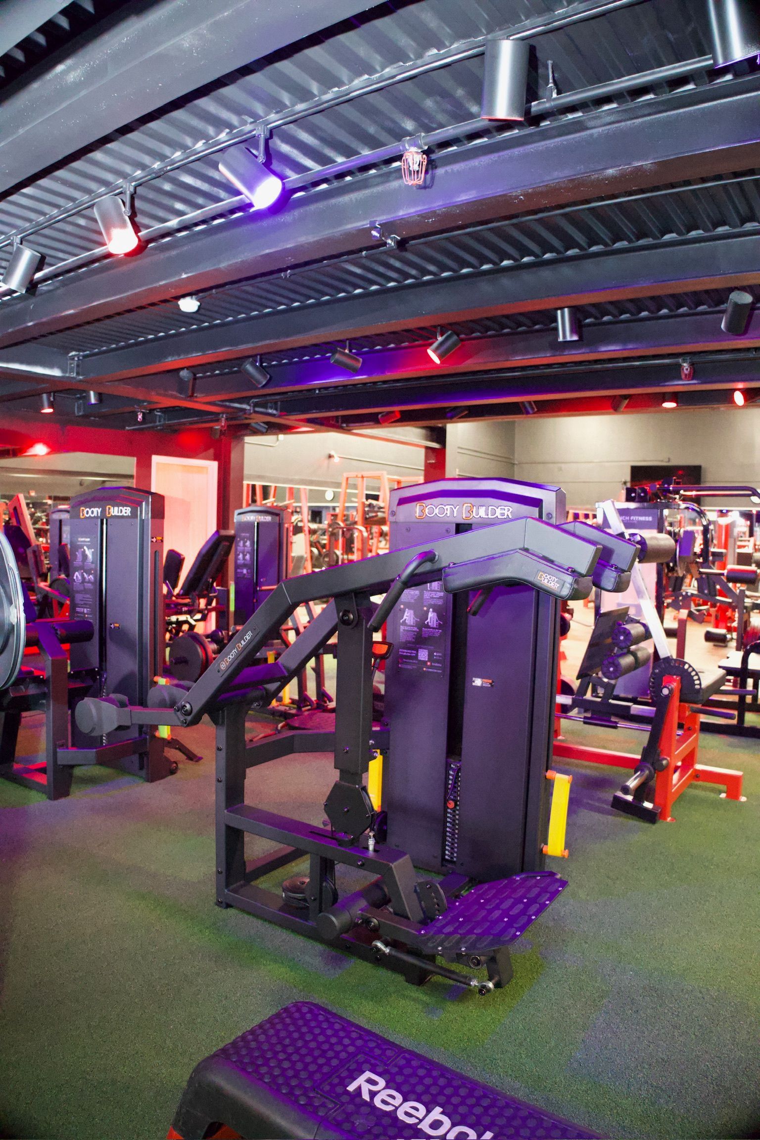 Gym interior with exercise machines, green floor, and red and purple lighting.