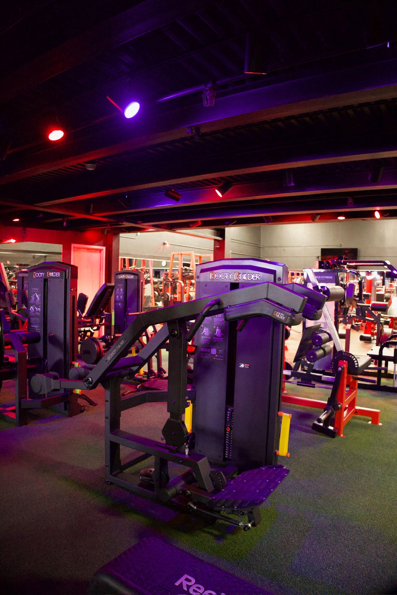 Gym interior with exercise machines, red and purple lighting, and green flooring.