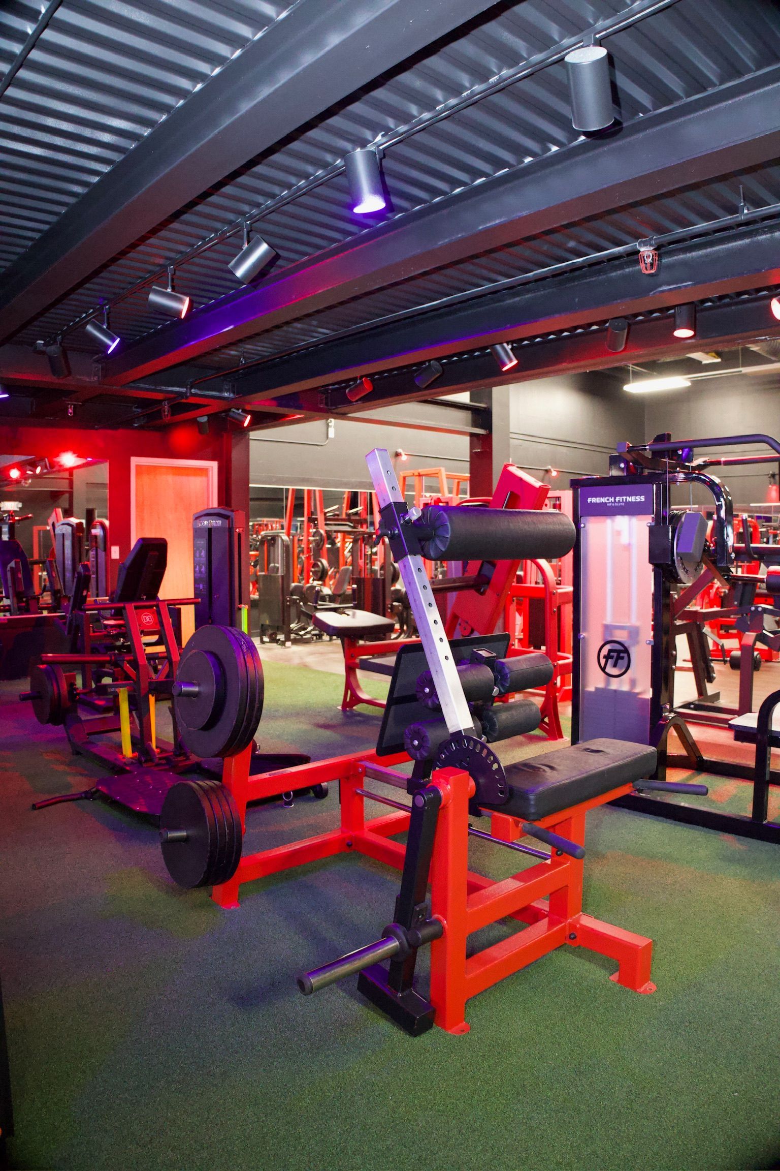 Gym interior with red and black exercise equipment on green flooring under a metal ceiling with track lighting.