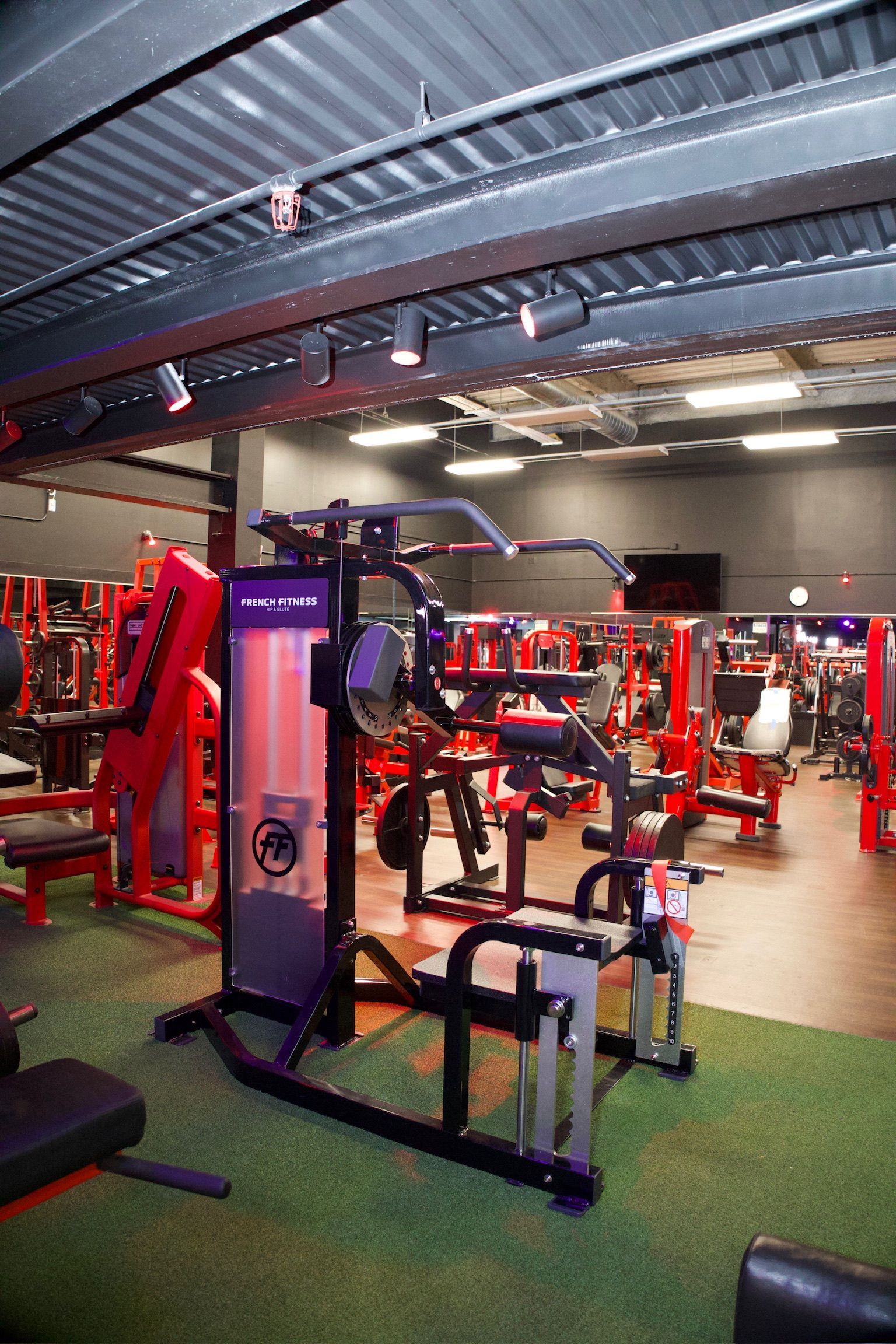 Gym interior with exercise machines, primarily red, set on green turf flooring. Black ceiling with lights.