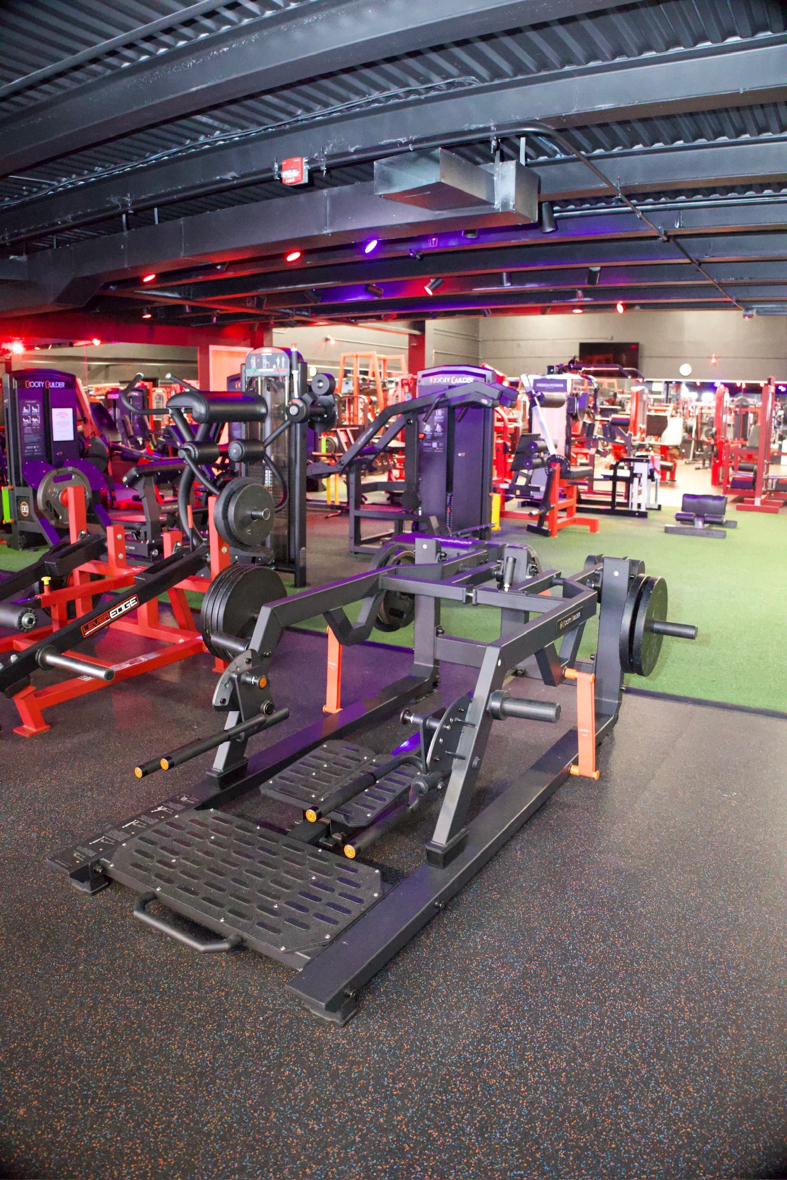 A weightlifting machine in a gym. Black and red equipment on a rubber floor. Other gym equipment is visible.