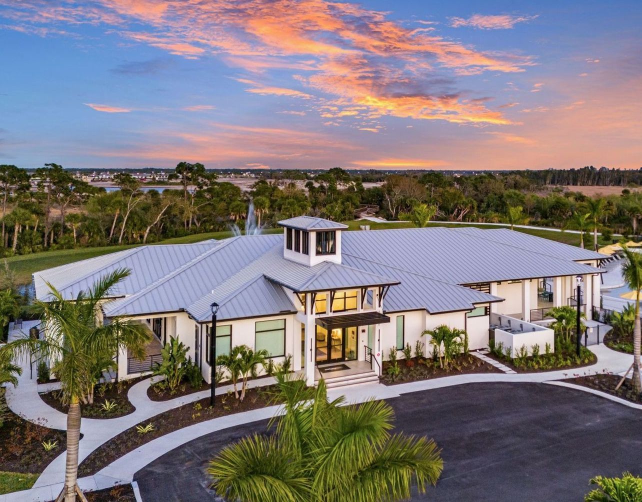 Exterior view of a modern building with a silver roof, white walls, and palm trees under a colorful sunset.