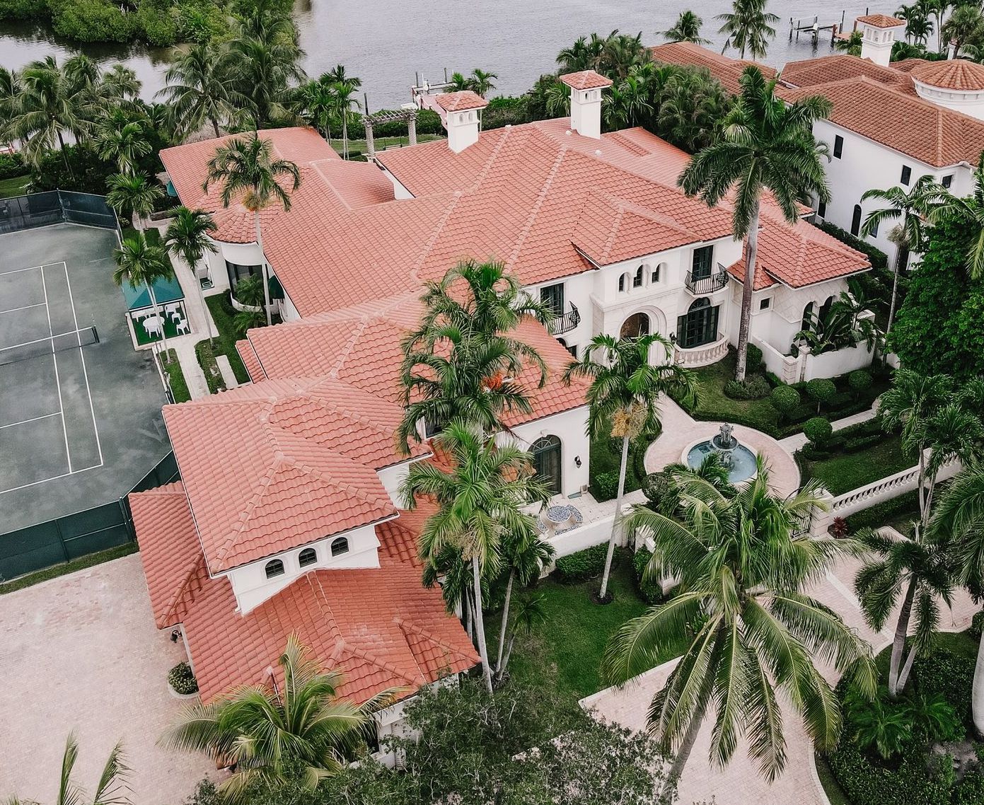 Luxury home with red tile roof, palm trees, and tennis court, near water.