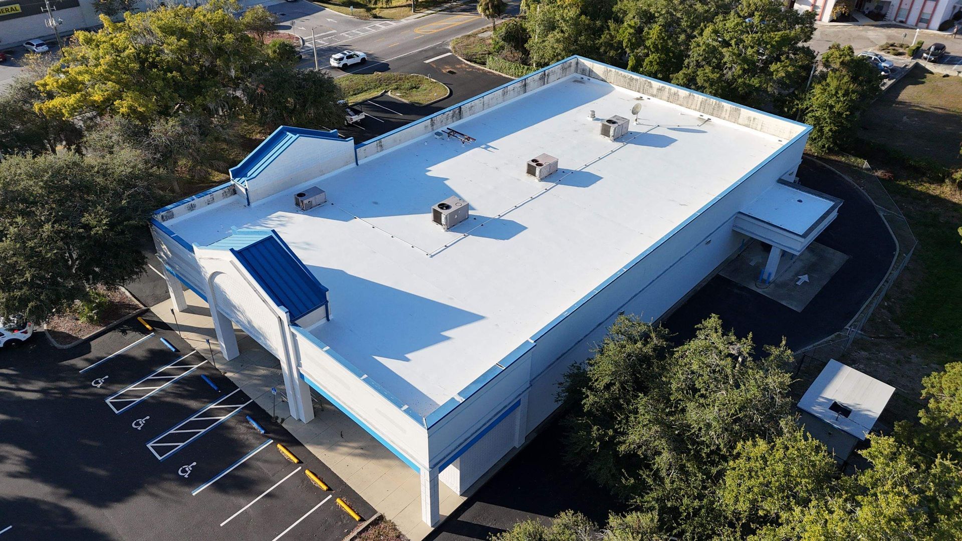 Aerial view of a white commercial building with a flat roof and air vents, surrounded by trees and parking.