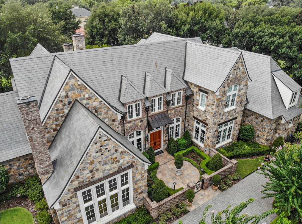 An aerial view of a large stone house with a gray roof surrounded by trees