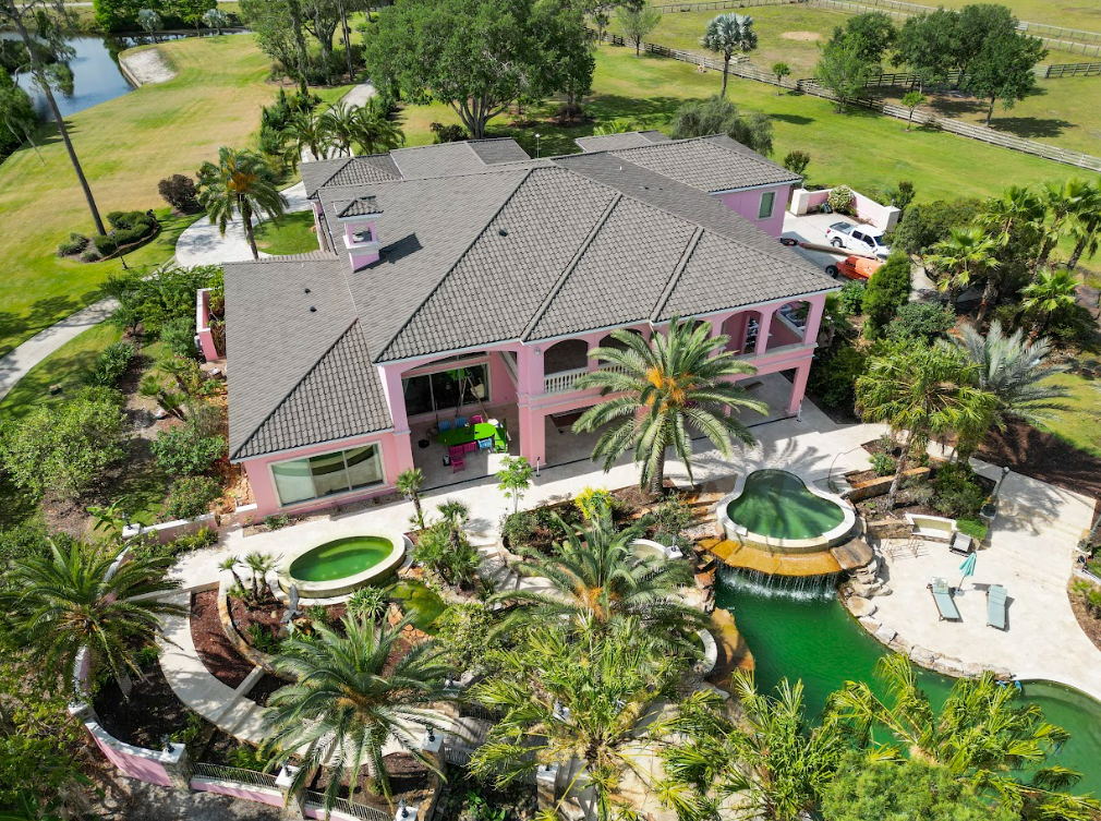 An aerial view of a large pink house surrounded by palm trees and a swimming pool