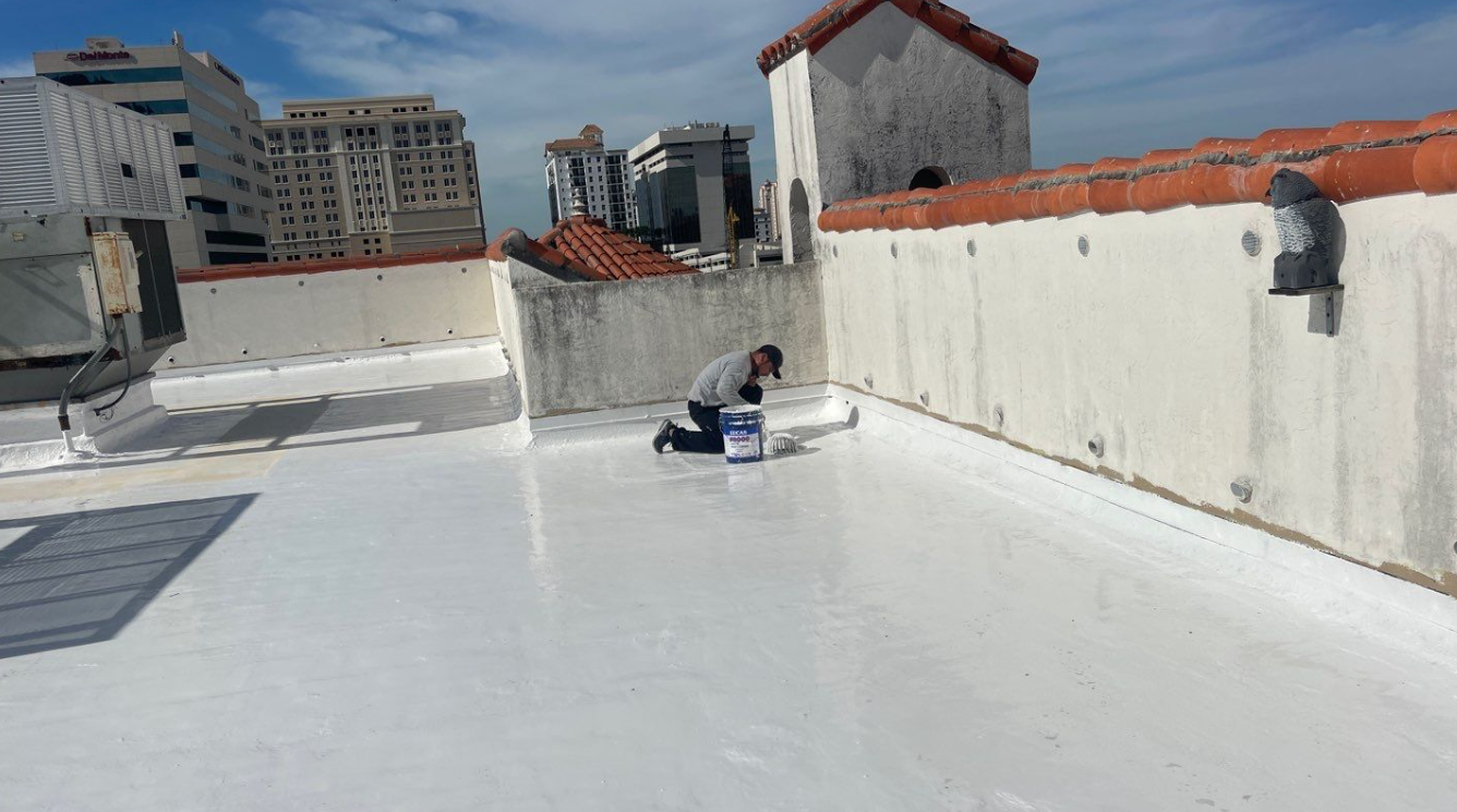 A man is kneeling on a roof painting it white