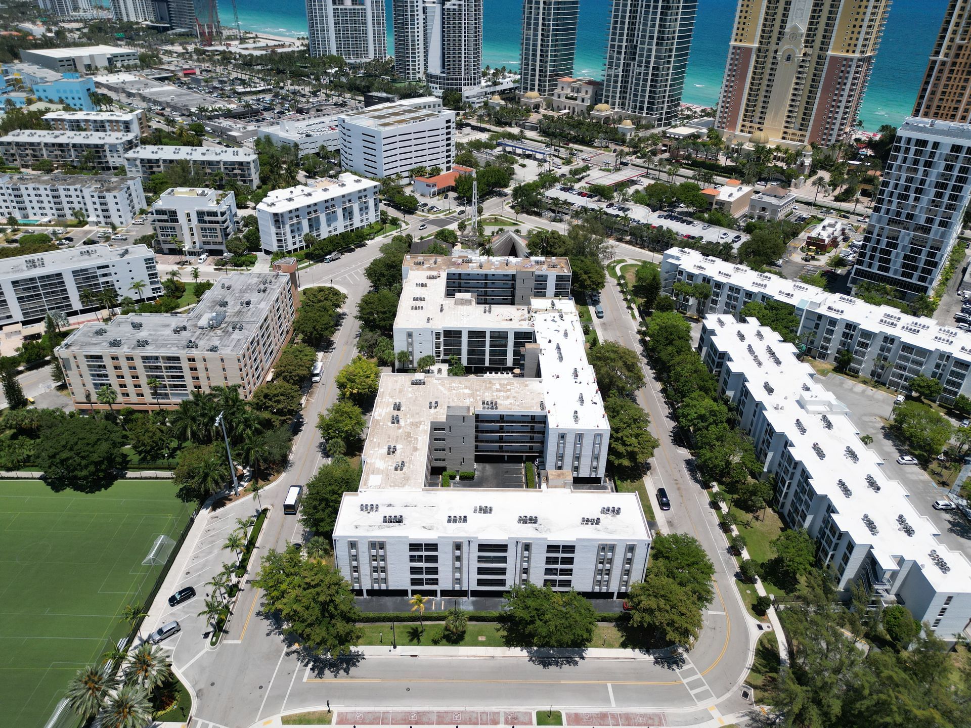 Aerial view of white apartment buildings surrounded by green trees and streets, with a blue ocean in the background.
