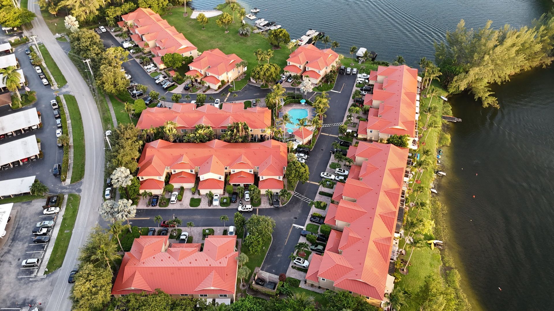 Aerial view of waterfront condos with red tile roofs, a pool, and parking; near a lake and trees.