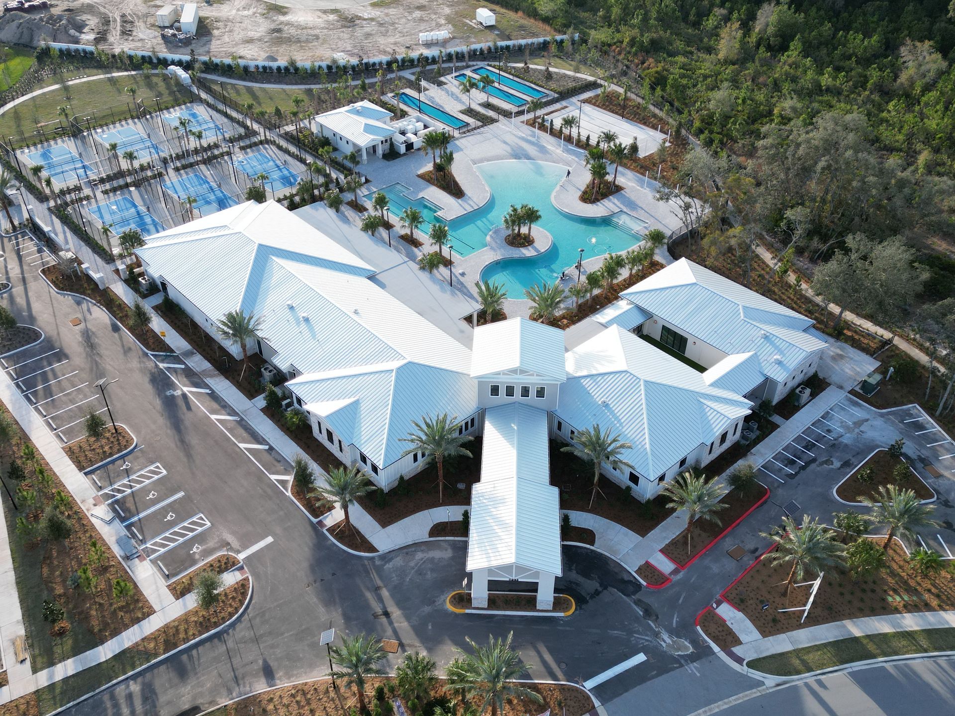 Aerial view of a white building complex with a pool, tennis courts, and parking lot.