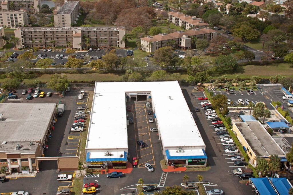 Aerial view of a U-shaped commercial building with cars, parking areas, and surrounding buildings under a sunny sky.