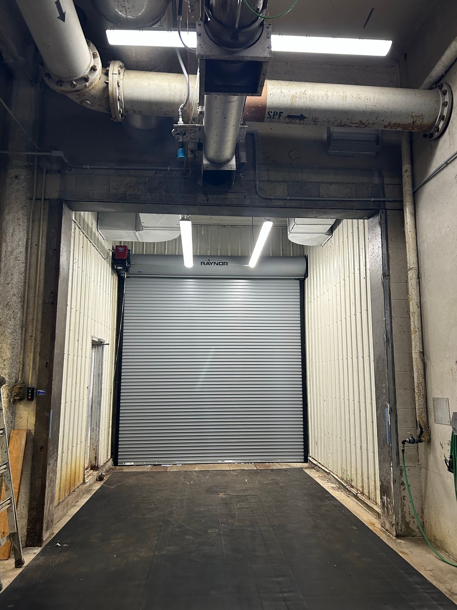 A garage door is open in a building with pipes hanging from the ceiling.
