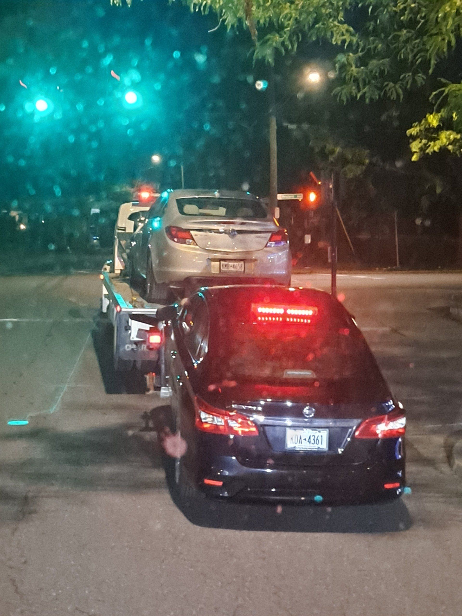 Two cars are being towed down a street at night.