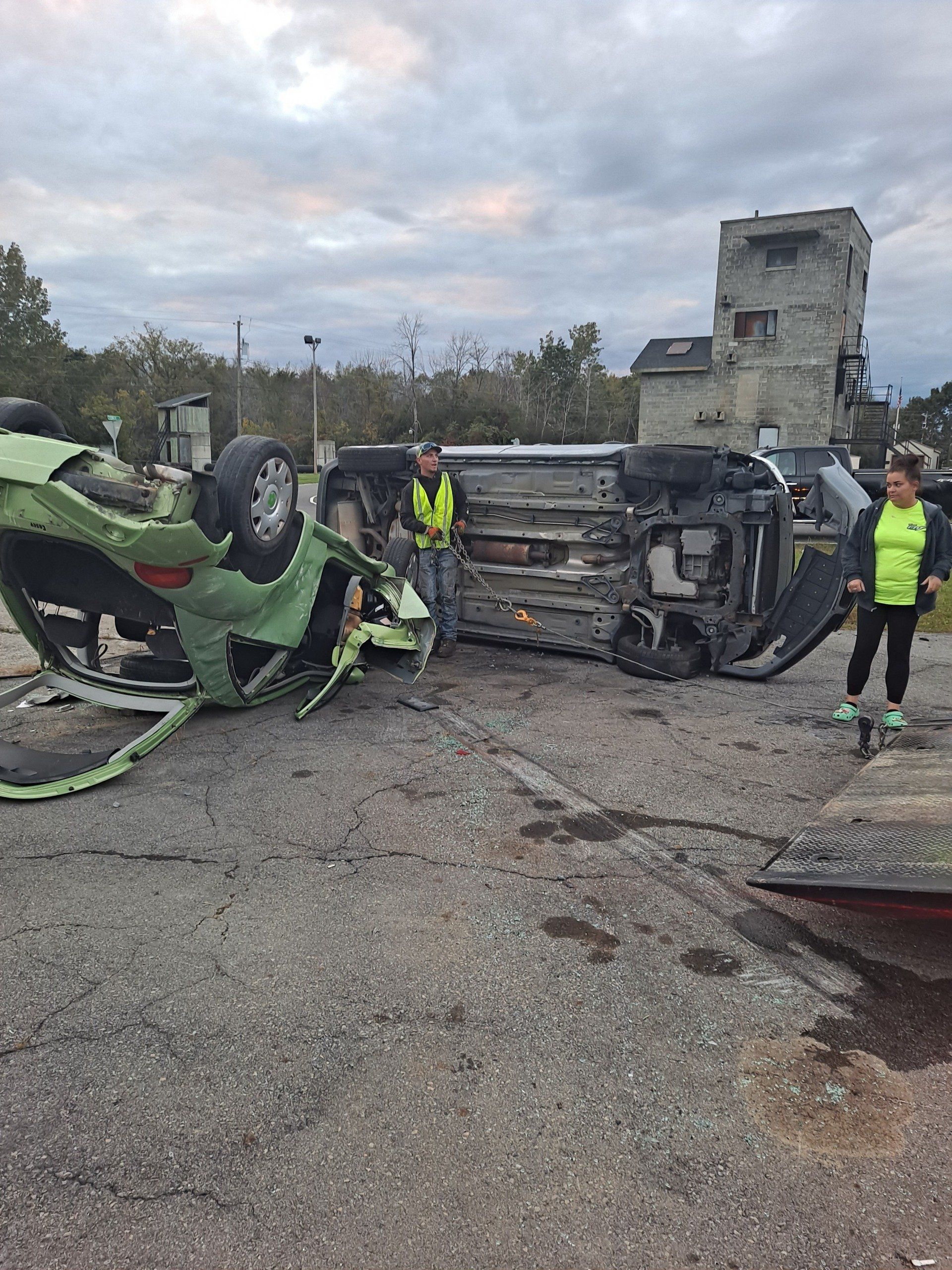 A man and a woman are standing next to a car that has turned on its side.