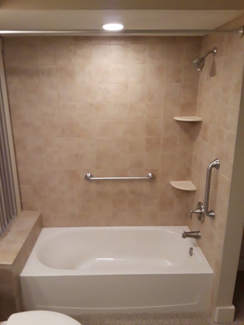 Empty beige tiled bathroom with a bathtub, grab bars, and shower shelves