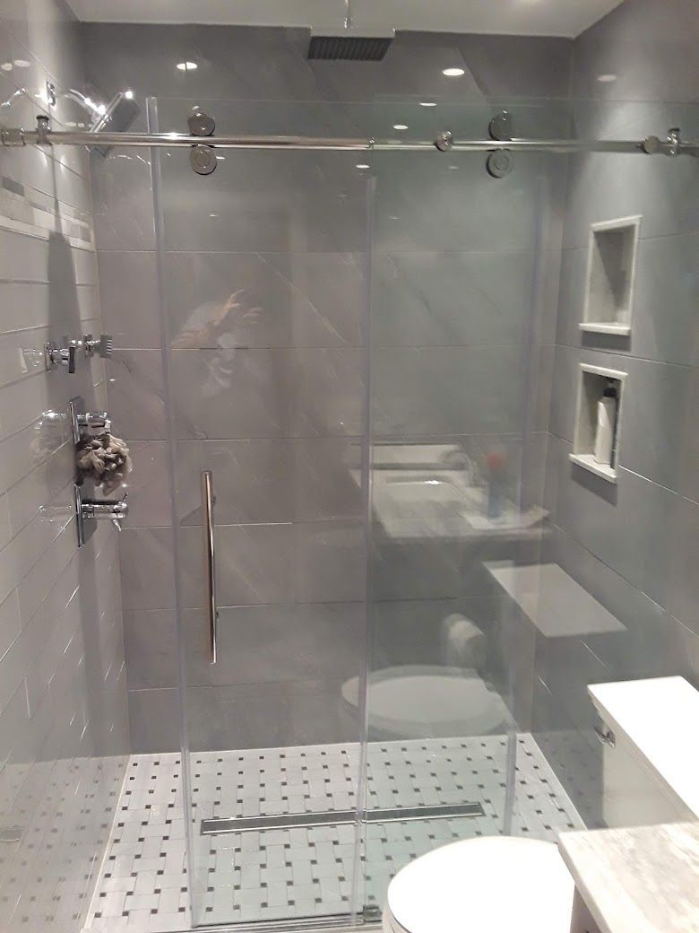 Modern bathroom with glass shower, gray tiles, white bench, and toilet visible behind the shower door