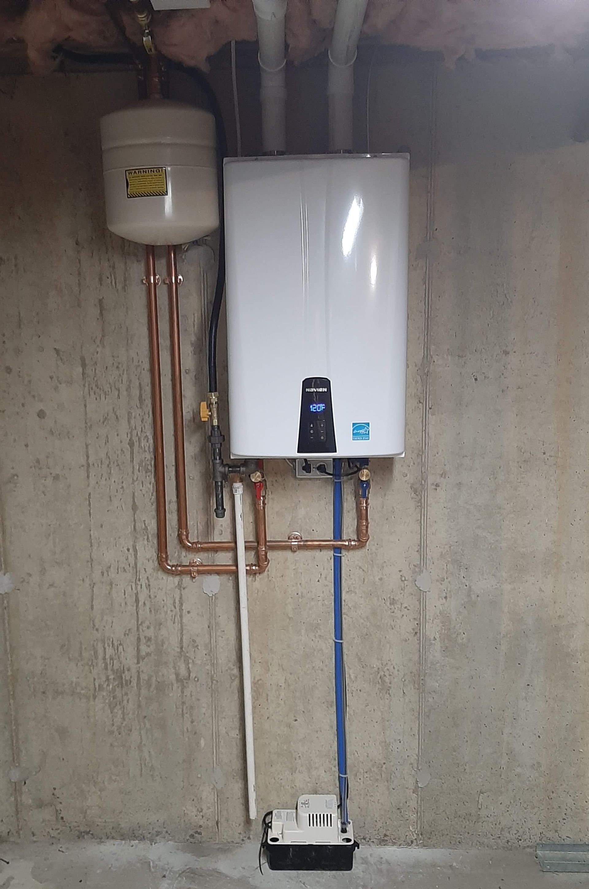Wall-mounted white water heater with copper pipes and a small tank in a concrete utility room