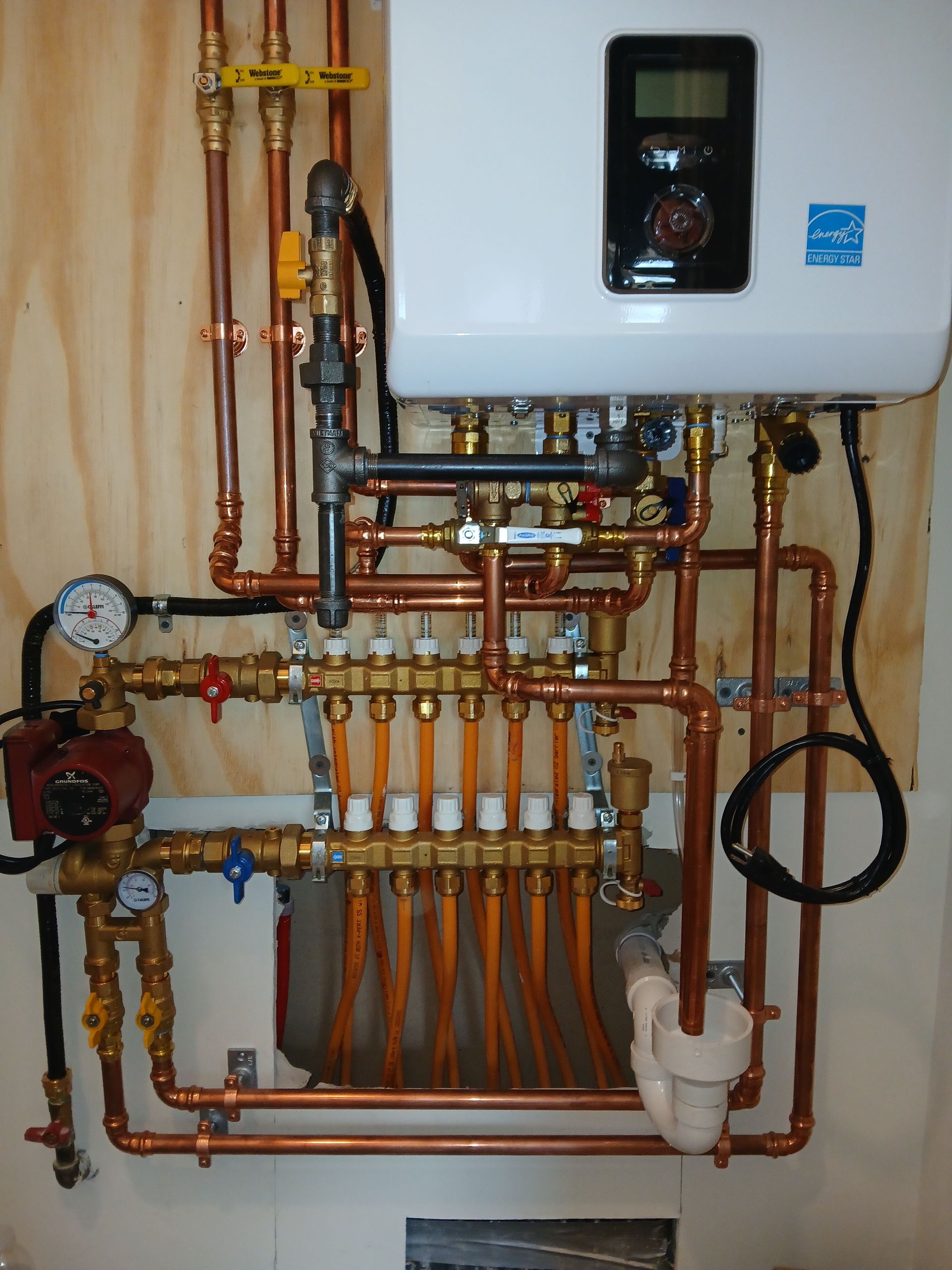 Wall-mounted boiler with copper pipes, valves, and white plumbing manifold in a utility room