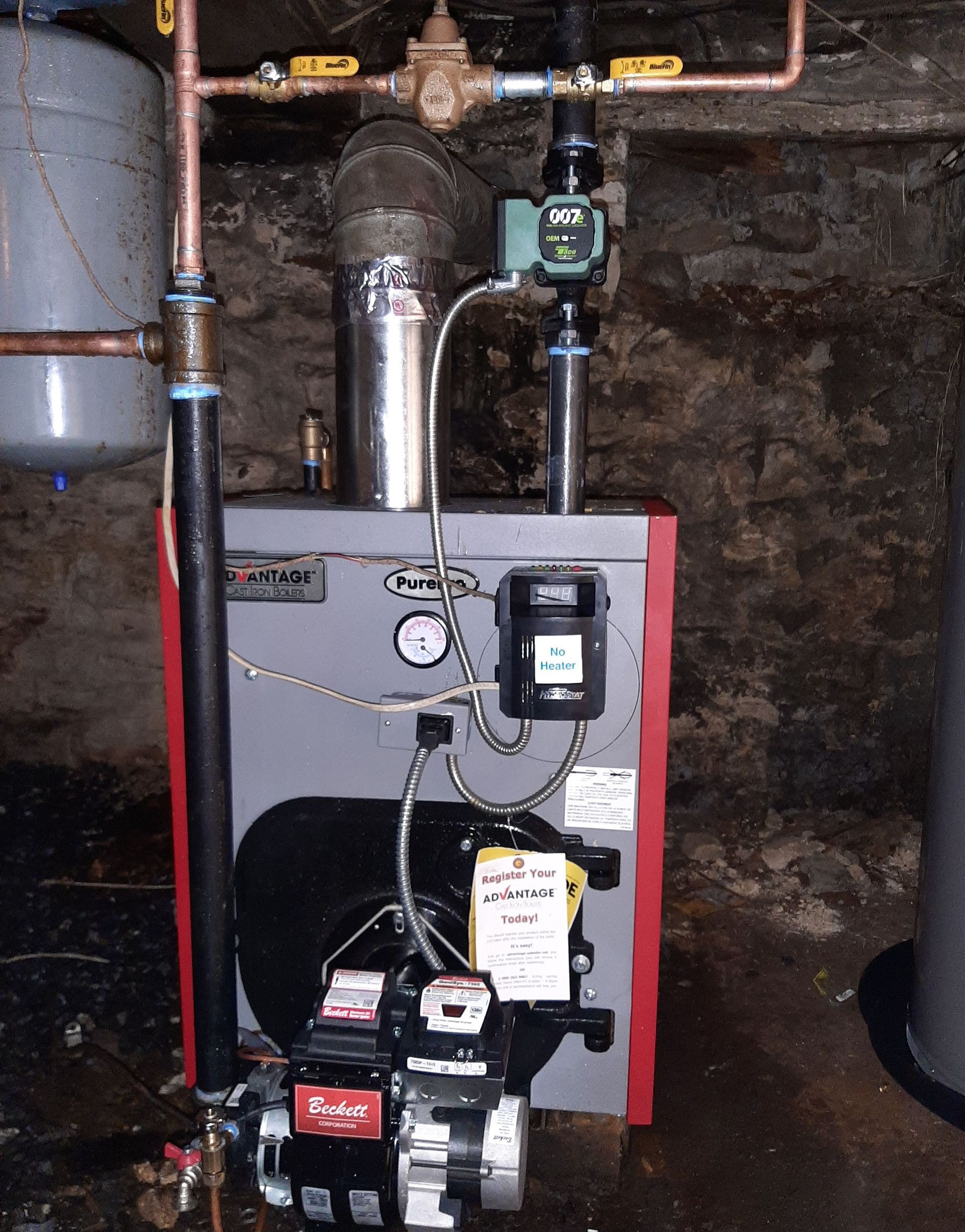 Boiler room with a gray furnace, red trim, pipes, gauges, and a pump assembly against a dark wall.