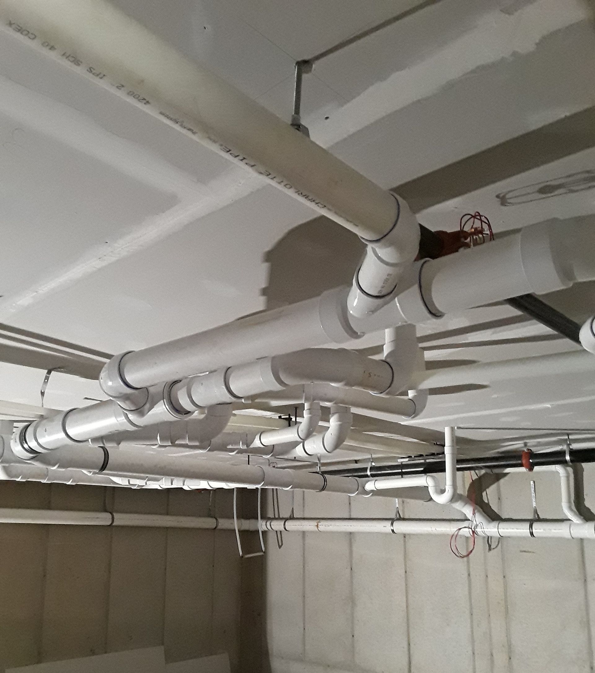 White PVC pipes and ceiling fixtures in an unfinished utility room with concrete walls and exposed ceiling.