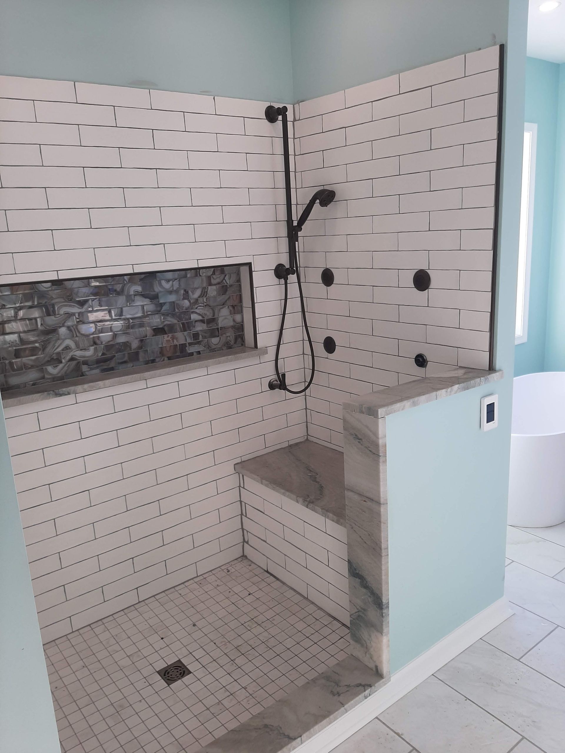 Modern bathroom shower with white tiled walls, gray bench, black fixtures, and frosted glass partition.